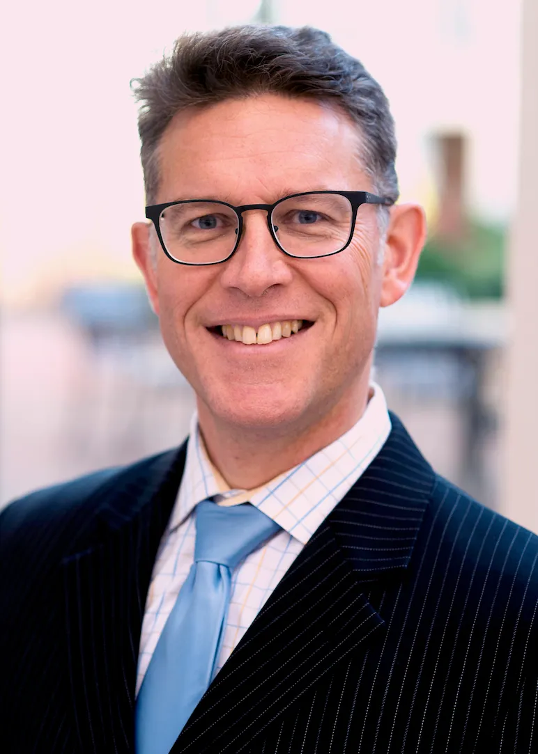 A headshot of Leamon. He is wearing a jacket and a blue tie, and smiling.