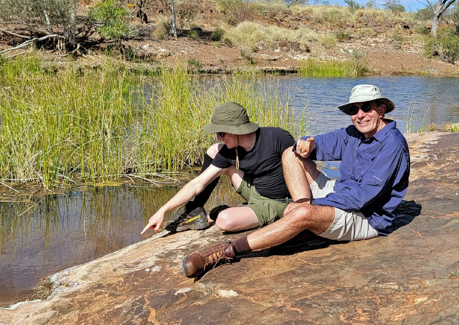 The researchers Dimitar Sasselov and Furkan Ozturk wearing hats and shorts, and sitting on the muddy shore of a body of water.