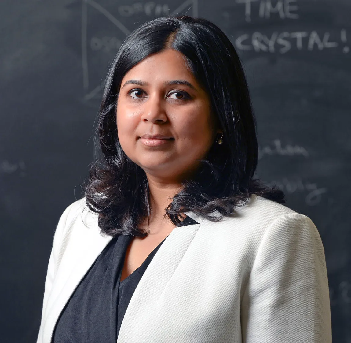 A portrait of Khemani wearing a white jacket and standing in front of a blackboard. The words “TIME CRYSTAL!” are visible on the board, along with some diagrams.