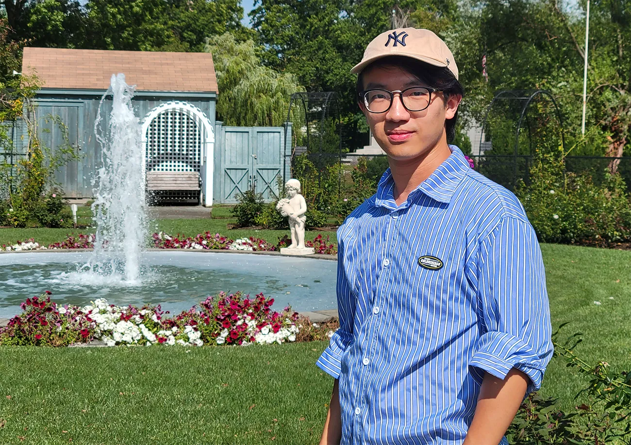 Ziming Liu stands in front of a fountain wearing a blue striped shirt and a baseball cap.