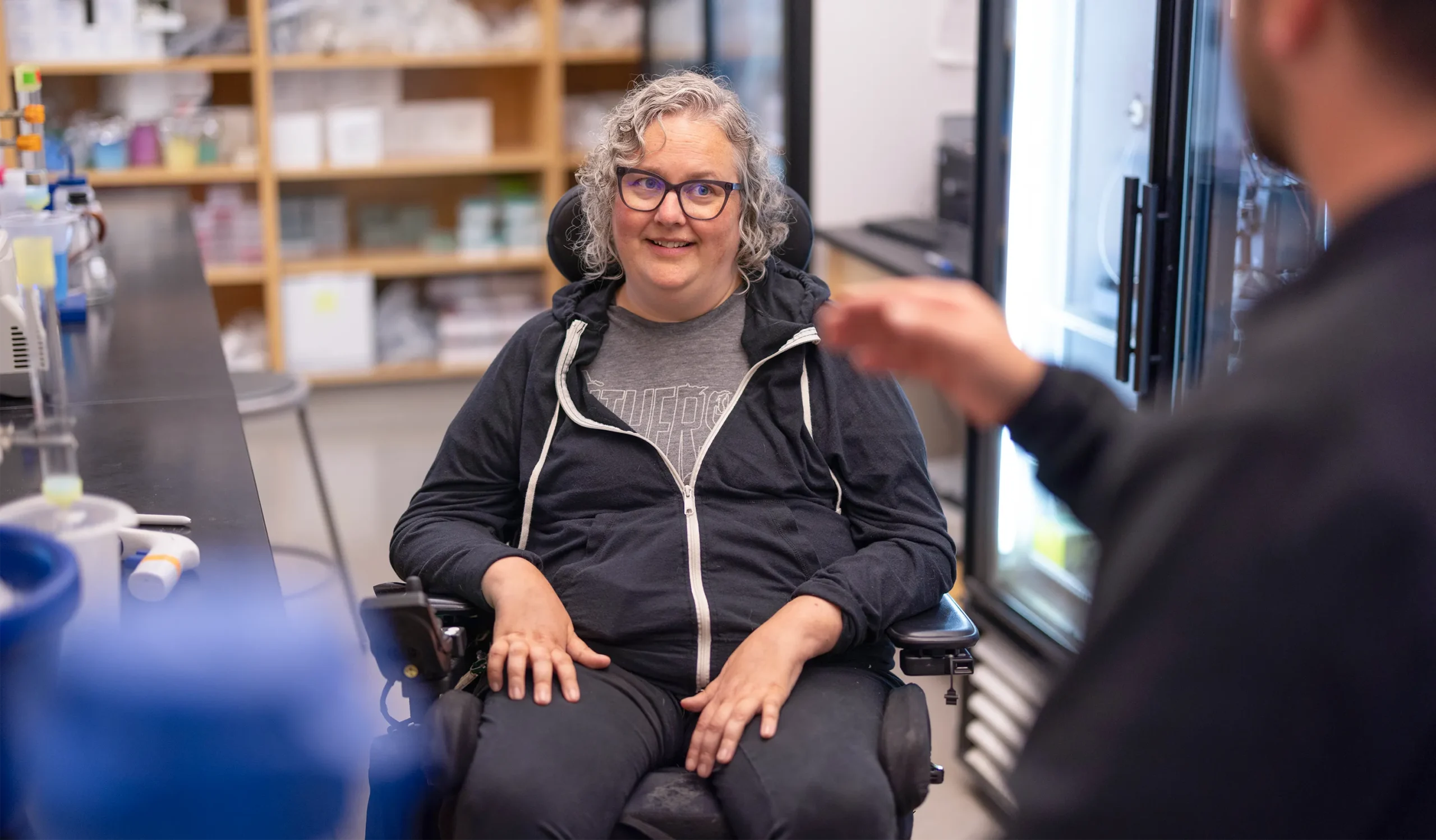 The researcher Carrie Partch sits in her motorized wheelchair in her laboratory, facing a coworker speaking and gesturing with one hand in the foreground of the photo.