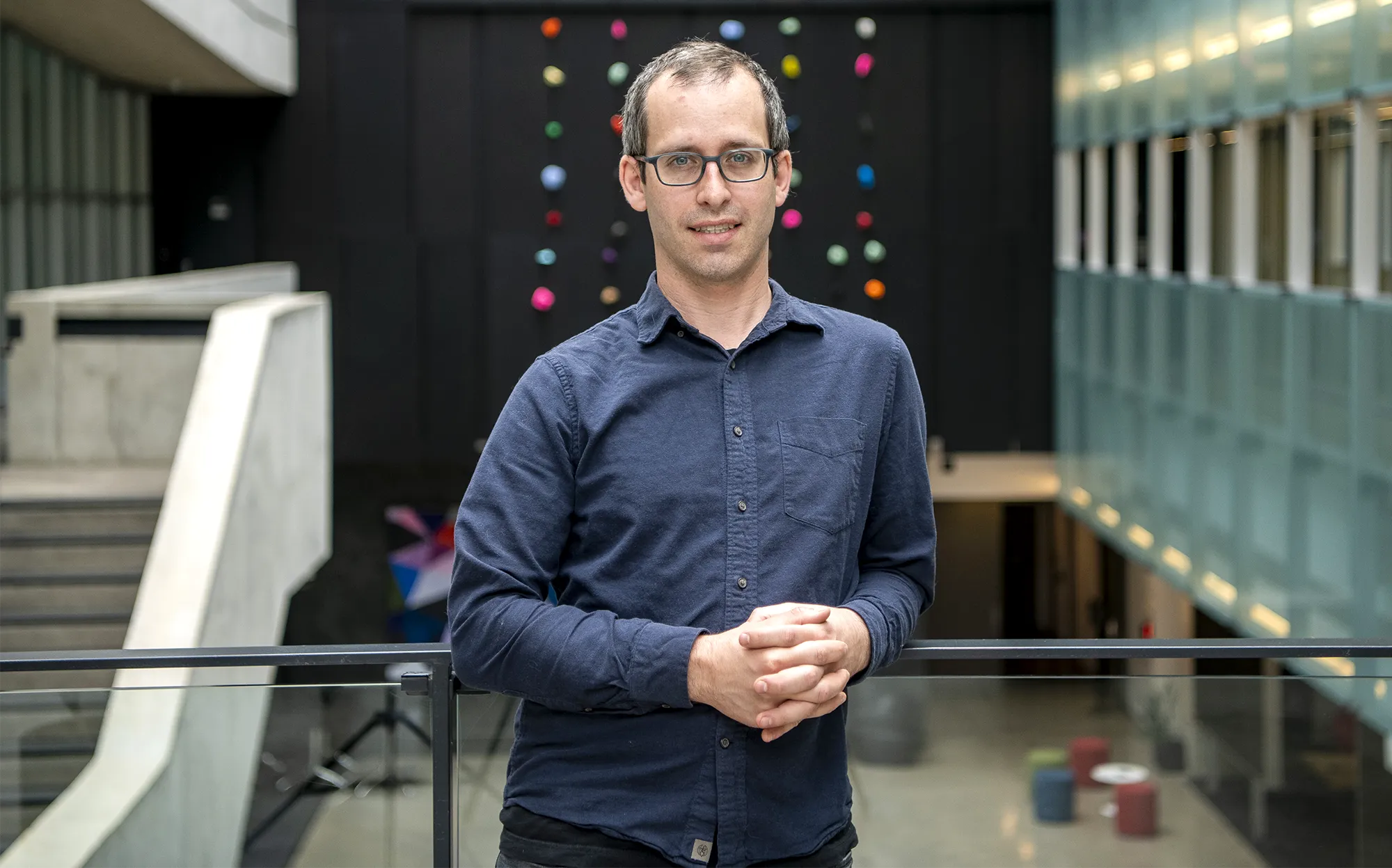 A man in a blue shirt stands with his hands clasped in front of him.