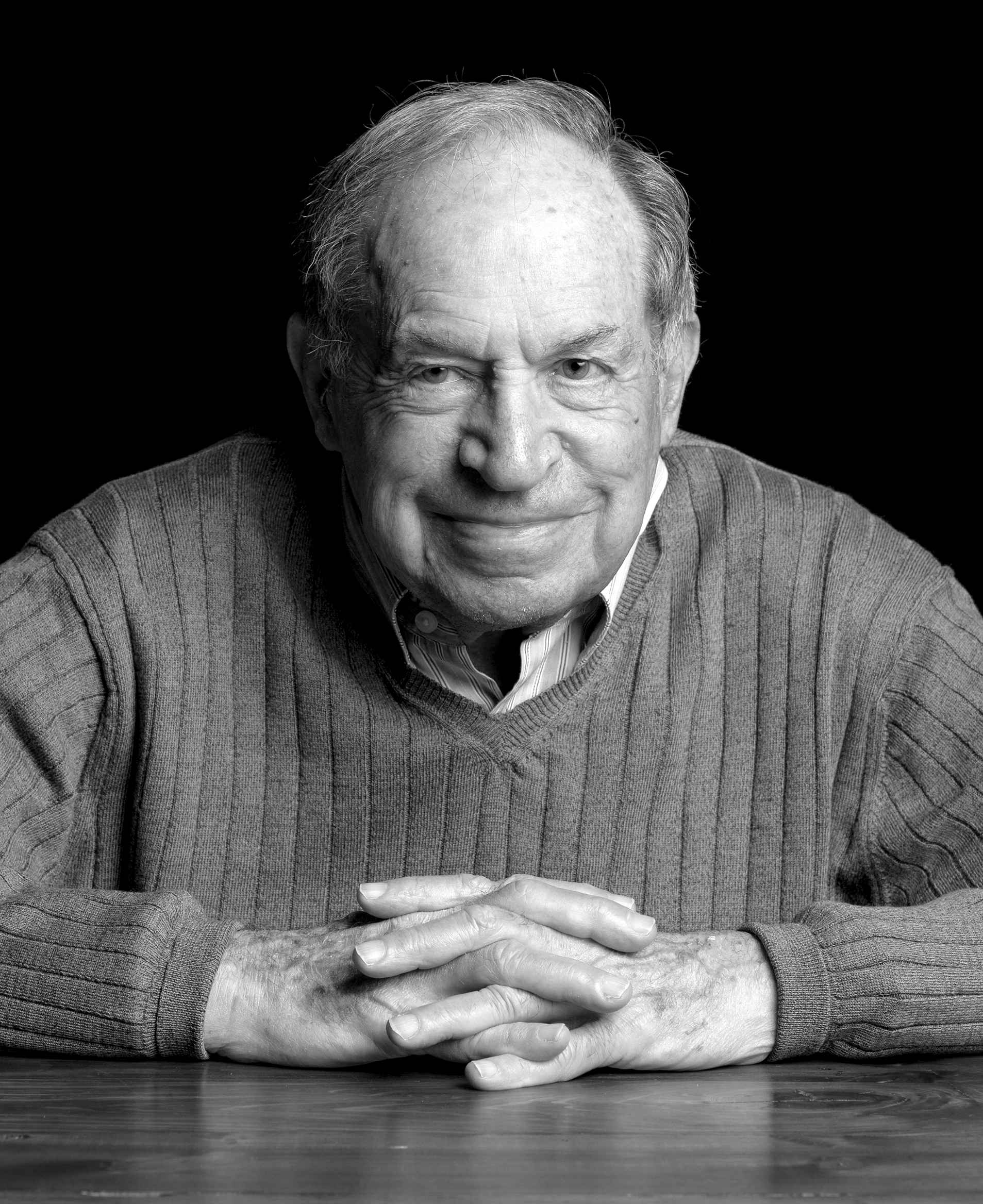 A man wearing a sweater leans forward over a table, with his hands folded.