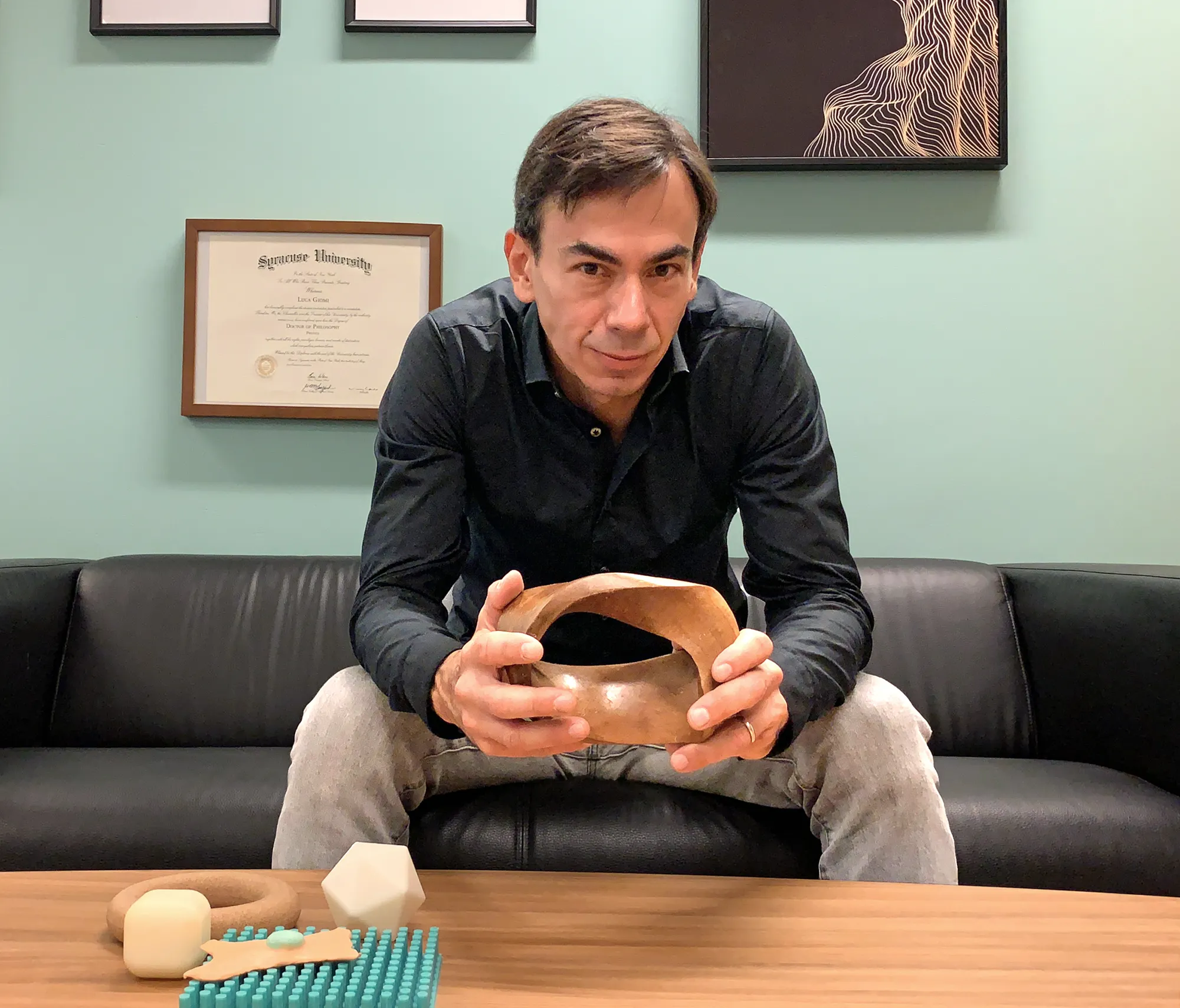 Giomi sitting on a couch, holding a geometric model. A diploma and some framed images are hanging on the wall behind him, which is painted a shade of aquamarine.