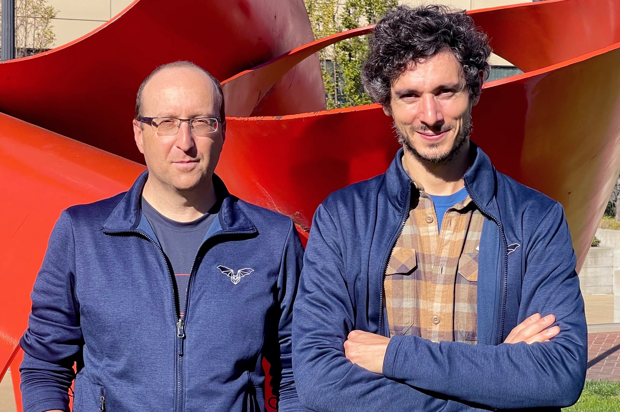 Michael Yartzev and Angelo Forli, both wearing jackets embroidered with an image of a bat, stand outside in front of a large red metal sculpture.