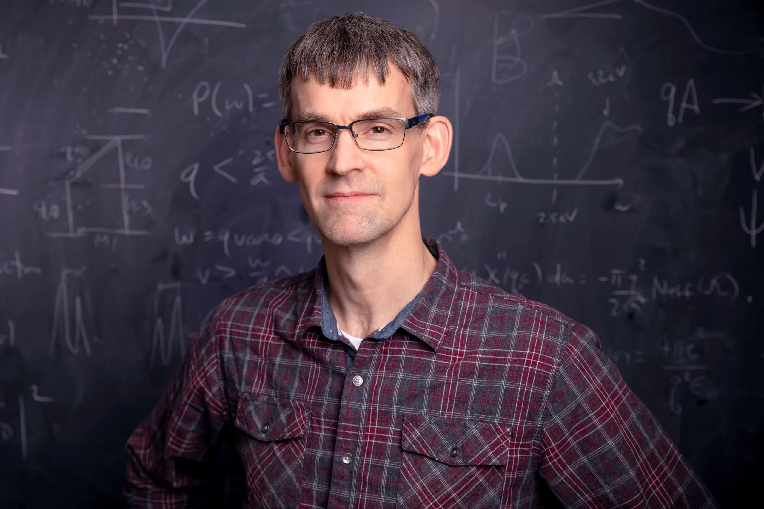 A bespectacled man stands in front of a blackboard covered in calculations.