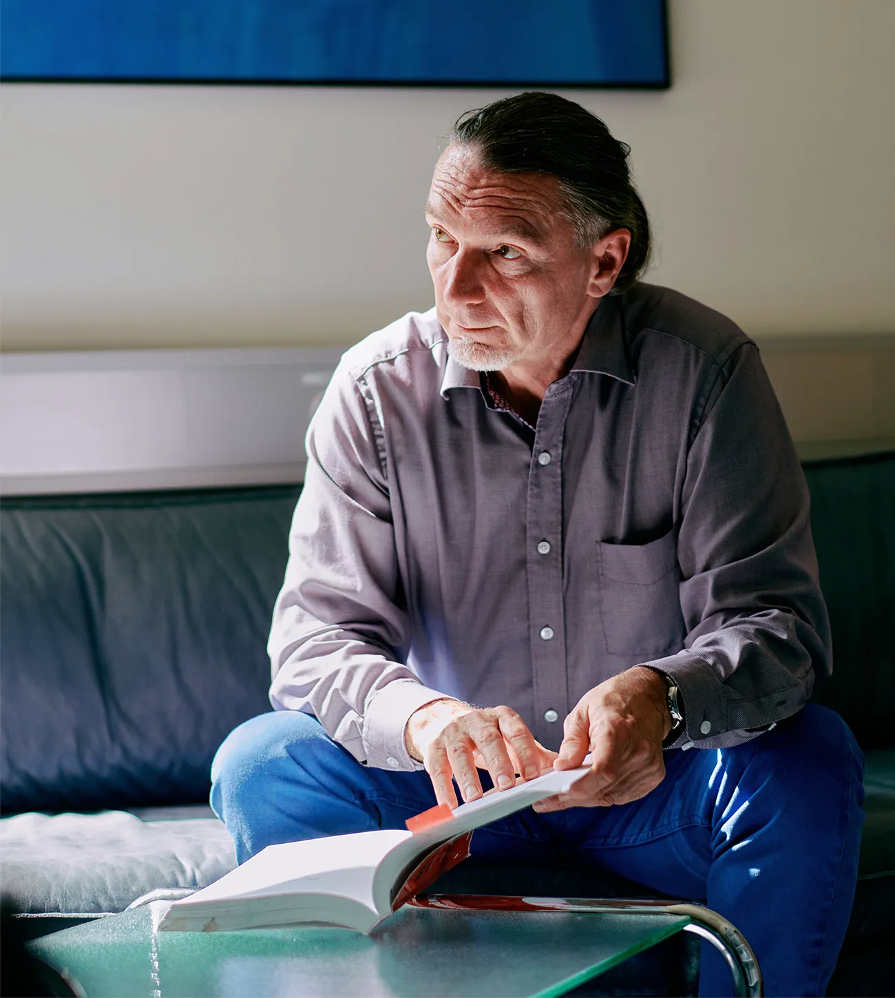 The biology researcher Andreas Wagner sits on a sofa, holding open a book that is on a glass table, while looking off to his right.