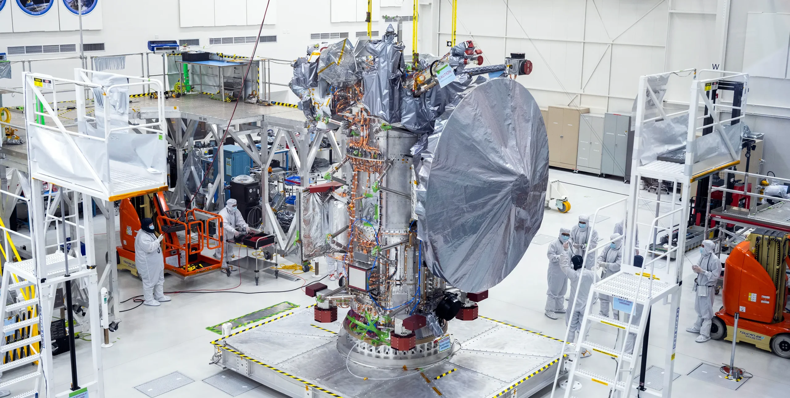 The Clipper spacecraft surrounded by technicians in clean suits in a clean room at the Jet Propulsion Laboratory. The spacecraft is tall and silvery, and not fully put together.