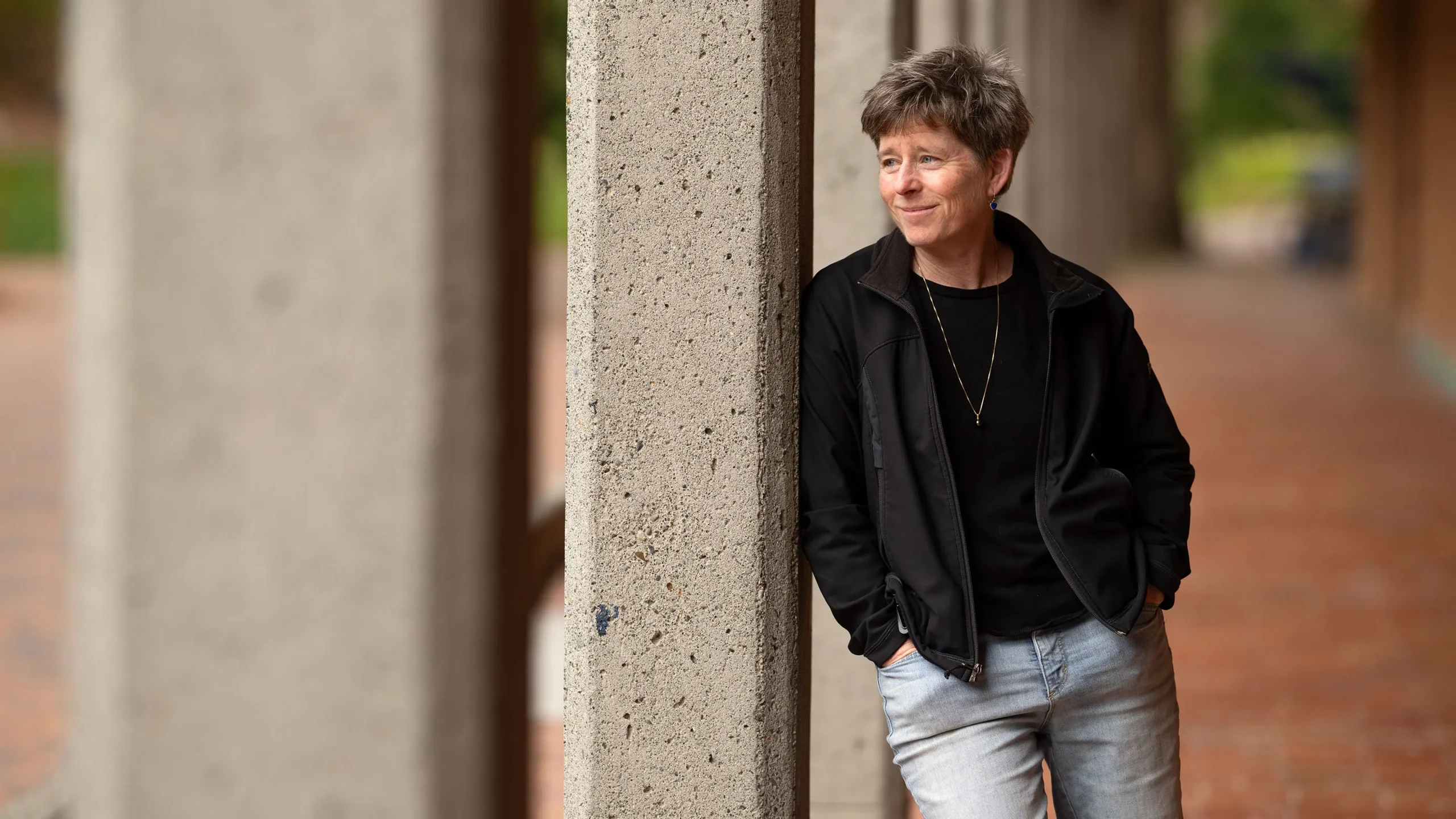 A portrait of Jackie Caplan-Auerbach. She is outside, leaning on a concrete pillar and staring into the distance.