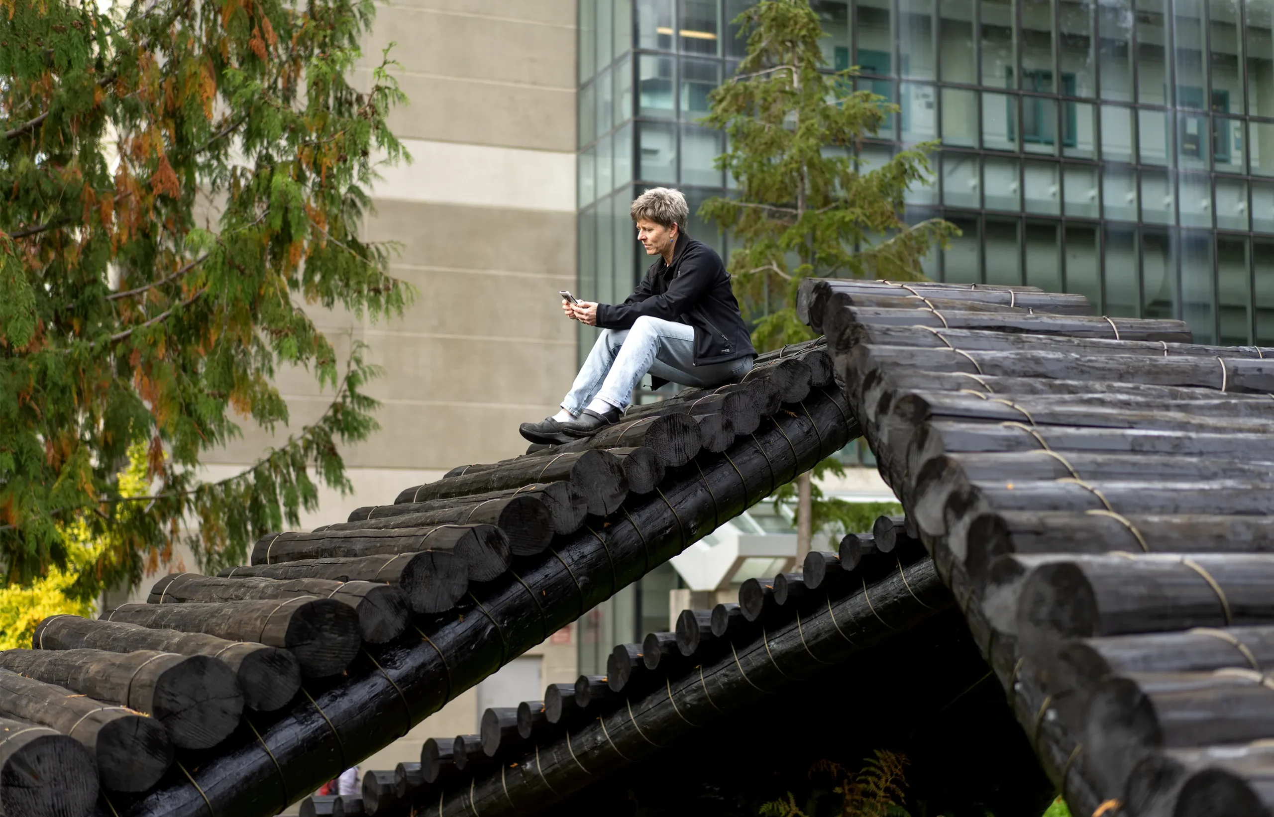 A portrait of Caplan-Auerbach on the Western Washington University campus. She is outside, sitting atop a structure made of logs, and staring at her cell phone.