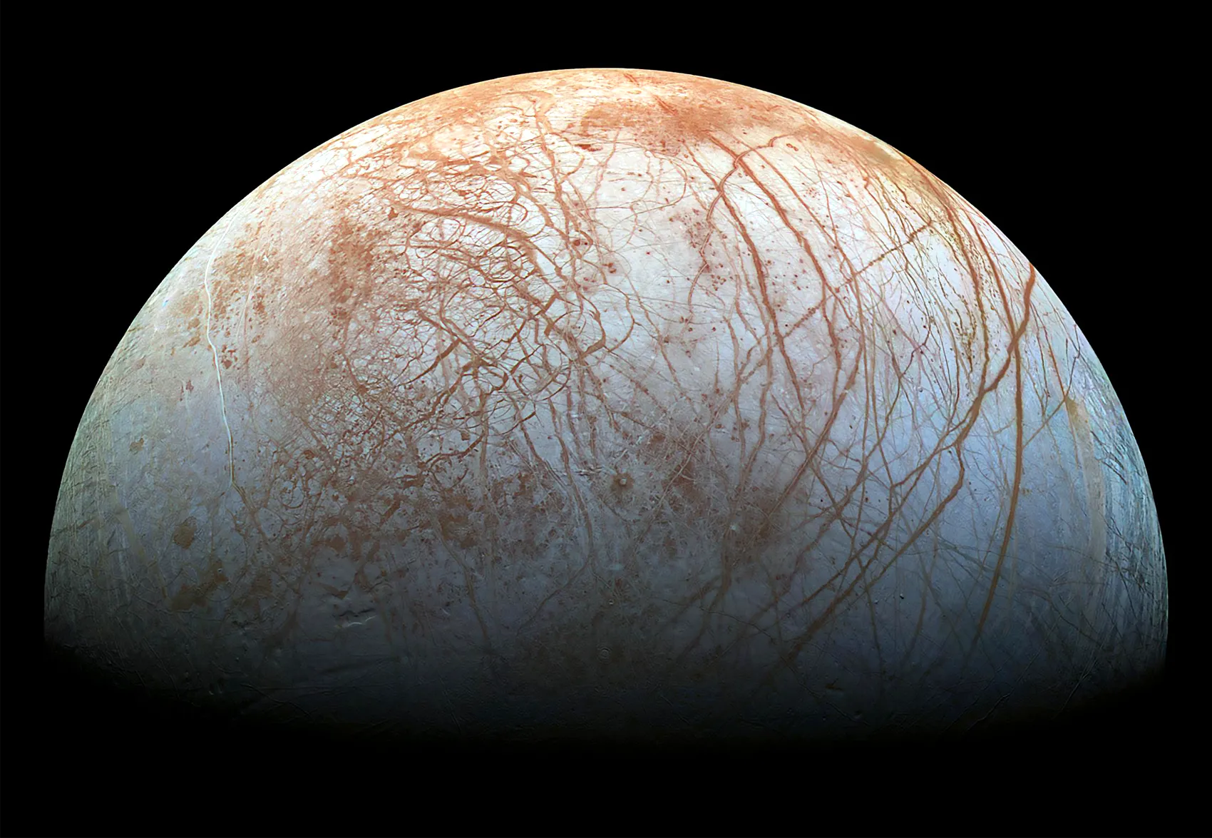 A photo of Europa made by the Galileo spacecraft. Half of the moon is lit up; the other half is in darkness. Europa’s surface is cracked and reddish.