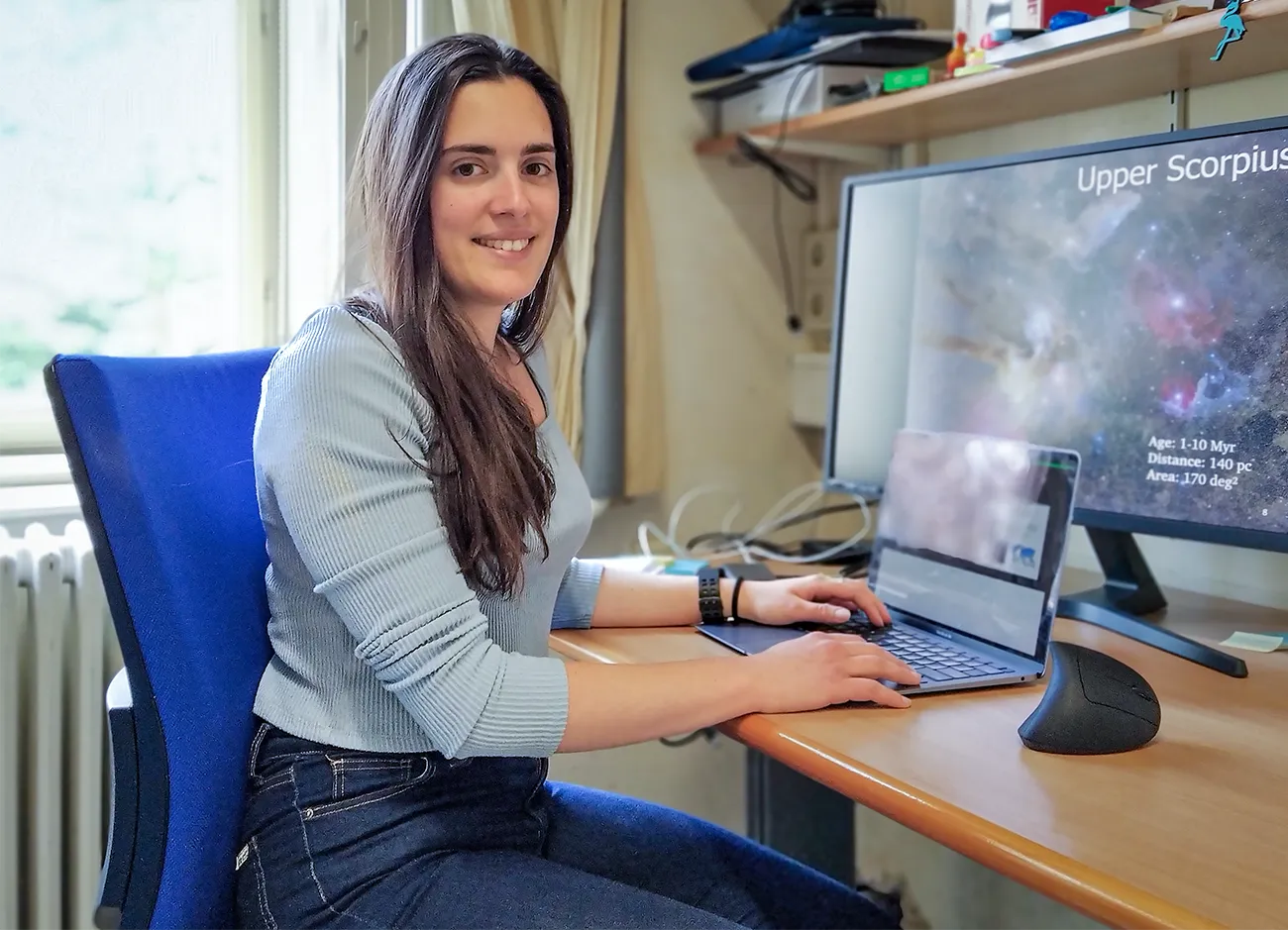 A portrait of Núria Miret Roig at her desk, facing the camera and smiling. On her computer screen is an image of the Scorpius nebula.