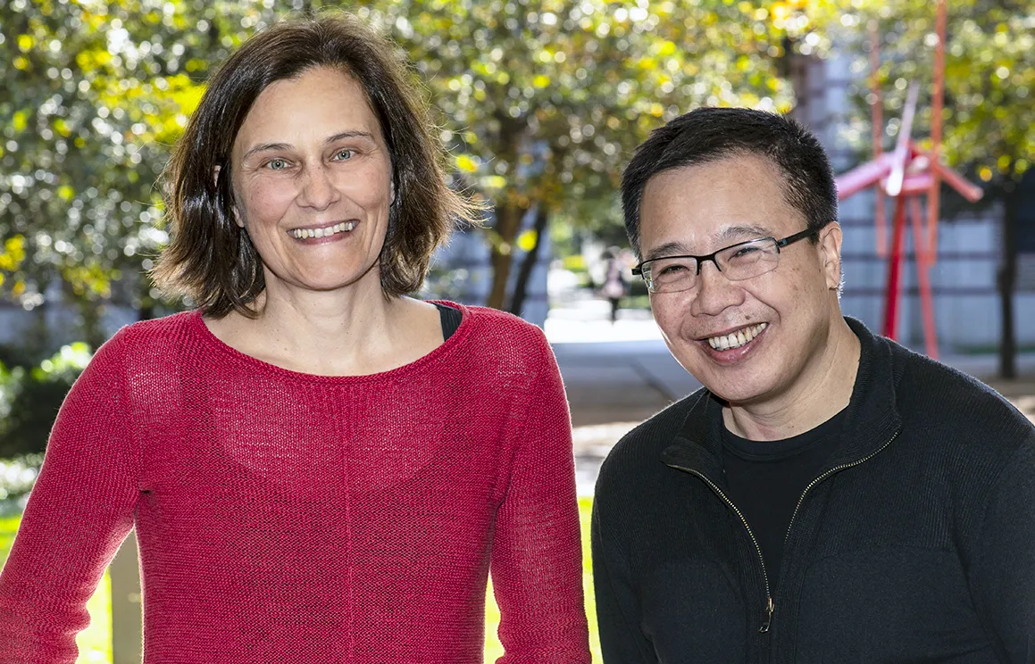 An image of two people. Silke Paschen is wearing a red sweater sits to the left of Qimiao Si, who wearing a black fleece. Leafy trees are behind them.