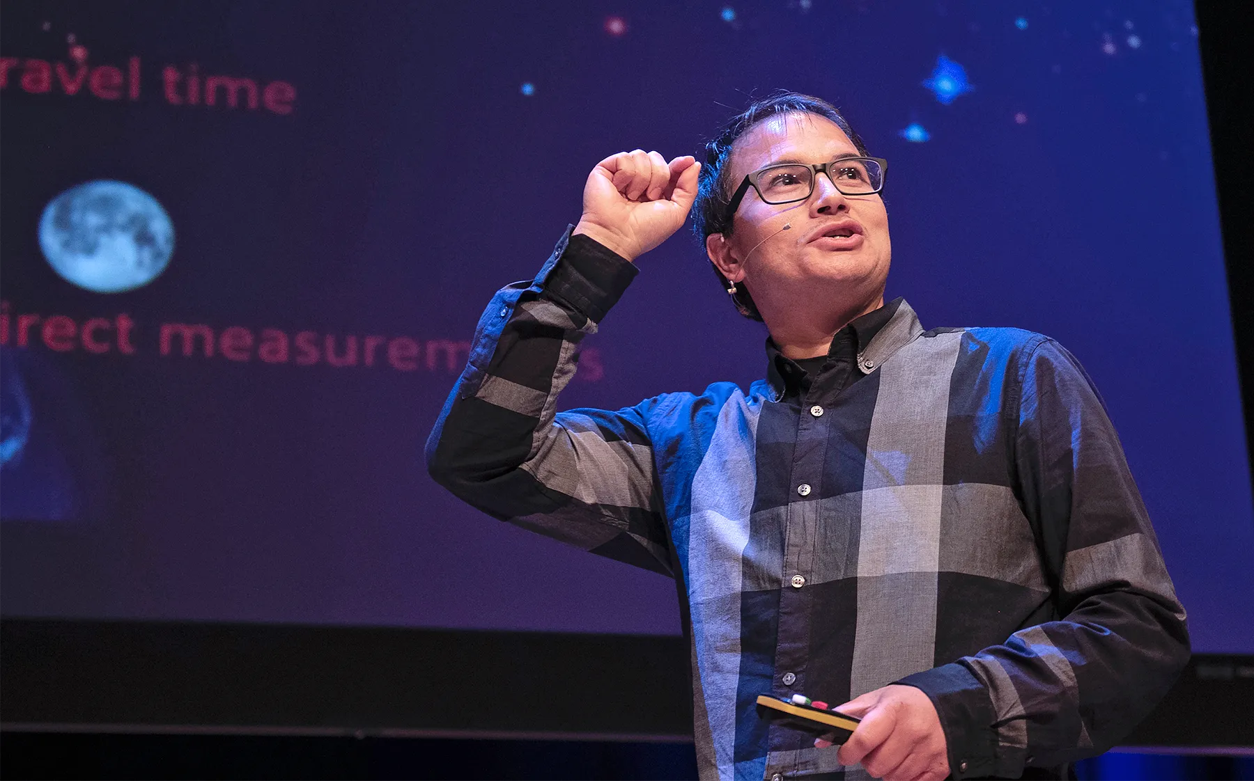 Andrew Levan wearing a plaid shirt and standing onstage, giving a public lecture.