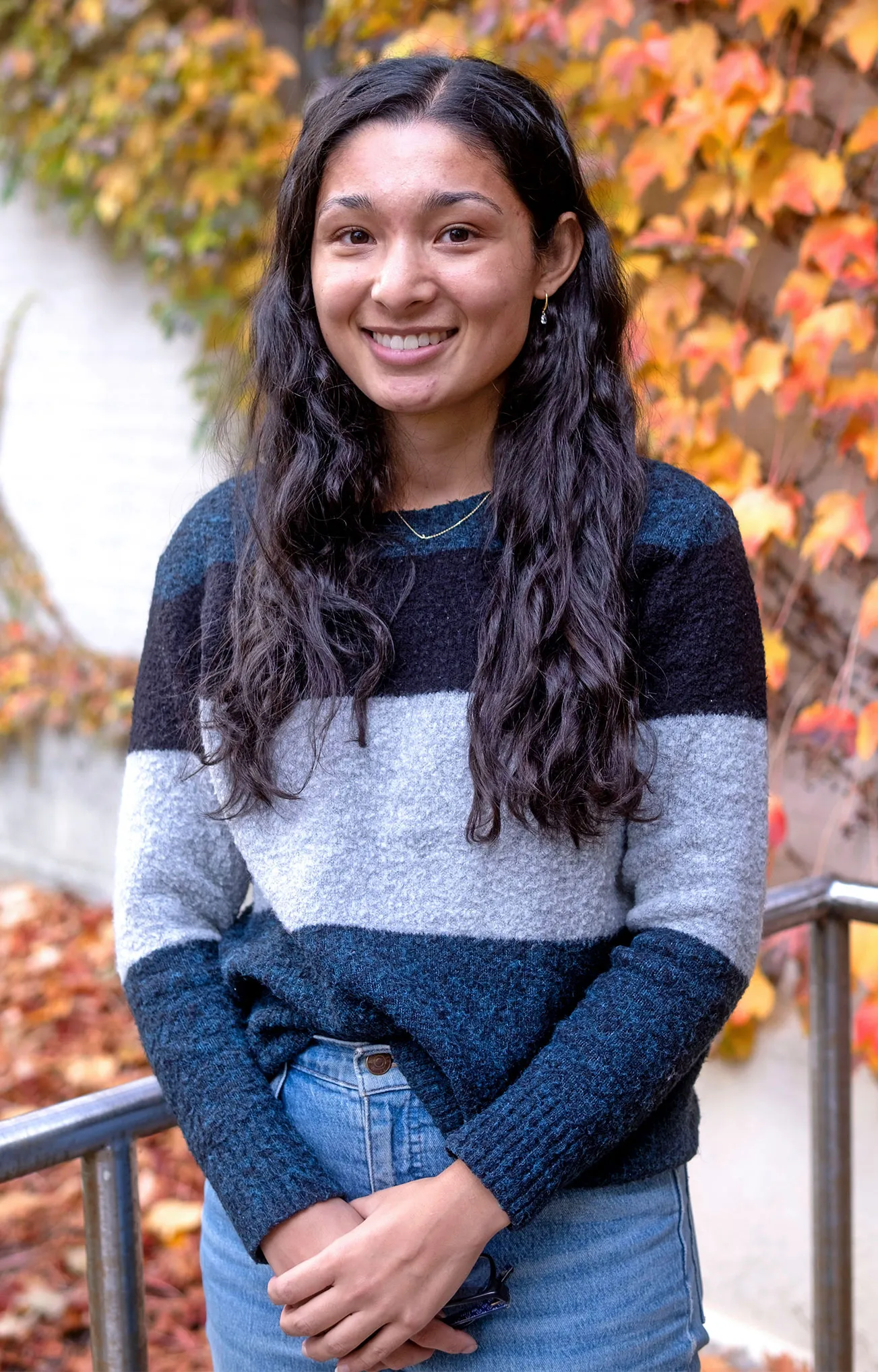 A portrait of Jillian Rastinejad. She is wearing a striped sweater and smiling. Behind her is a wall covered in autumn leaves.