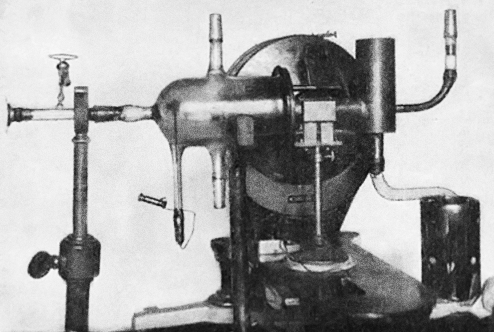 A black and white photograph of a complex contraption that sits on a table. It has multiple chambers connected by tubes and all are enclosed in a vacuum chamber.