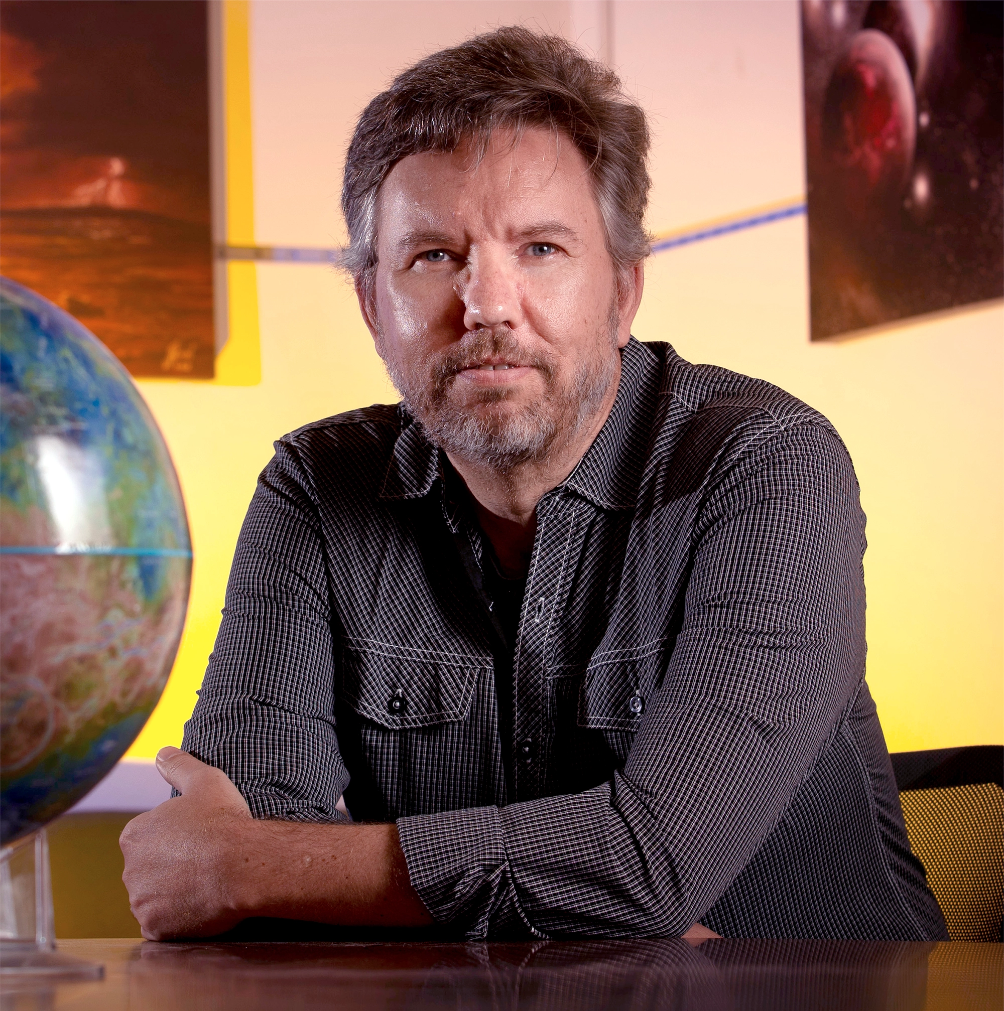 A portrait of Stephen Kane. His arms are crossed and he looks thoughtful. Behind him, a soft yellow glow illuminates a wall with images of planets.