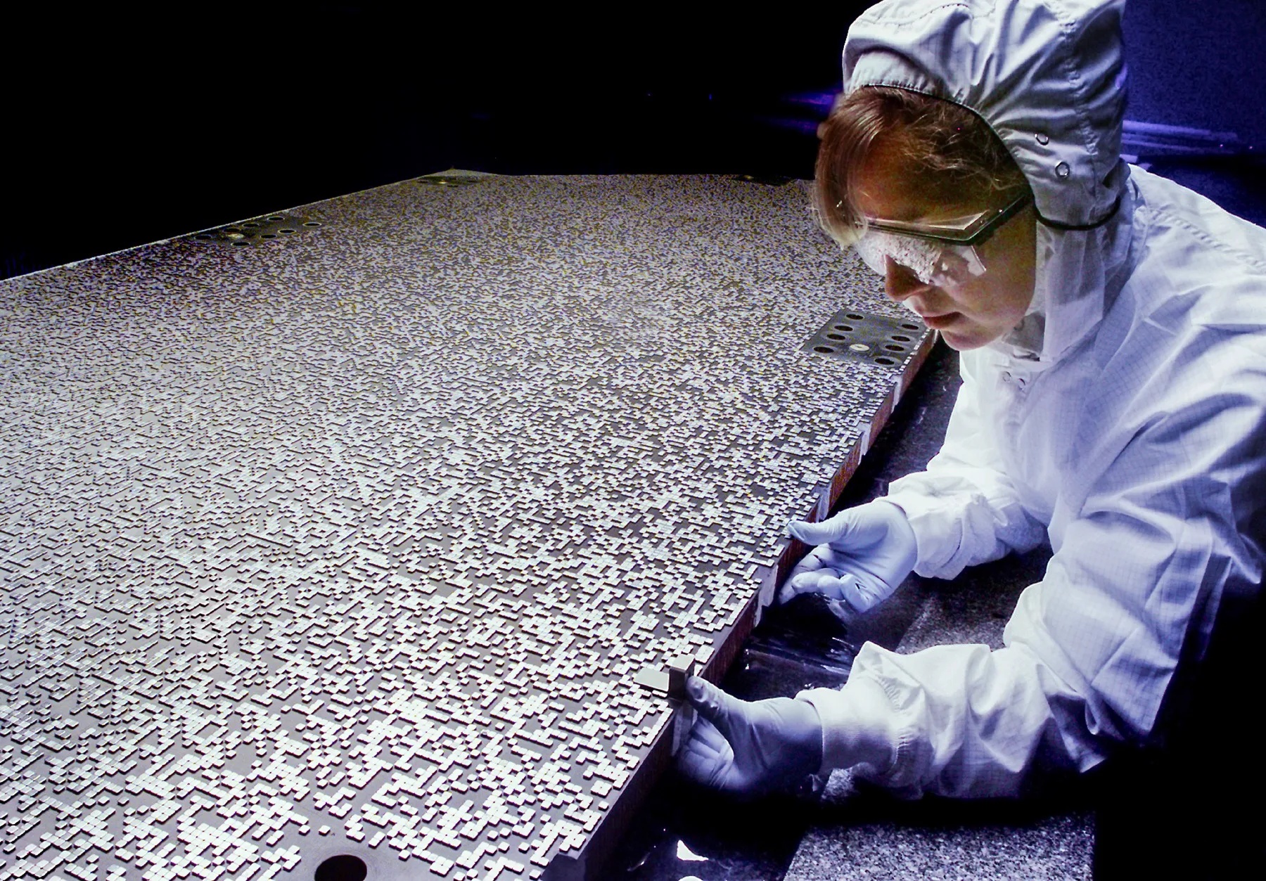 A technician in a clean suit fiddles with a lead plate the size of a dinner table. The plate is covered in patterns, made from squares, that collectively resemble a QR code.