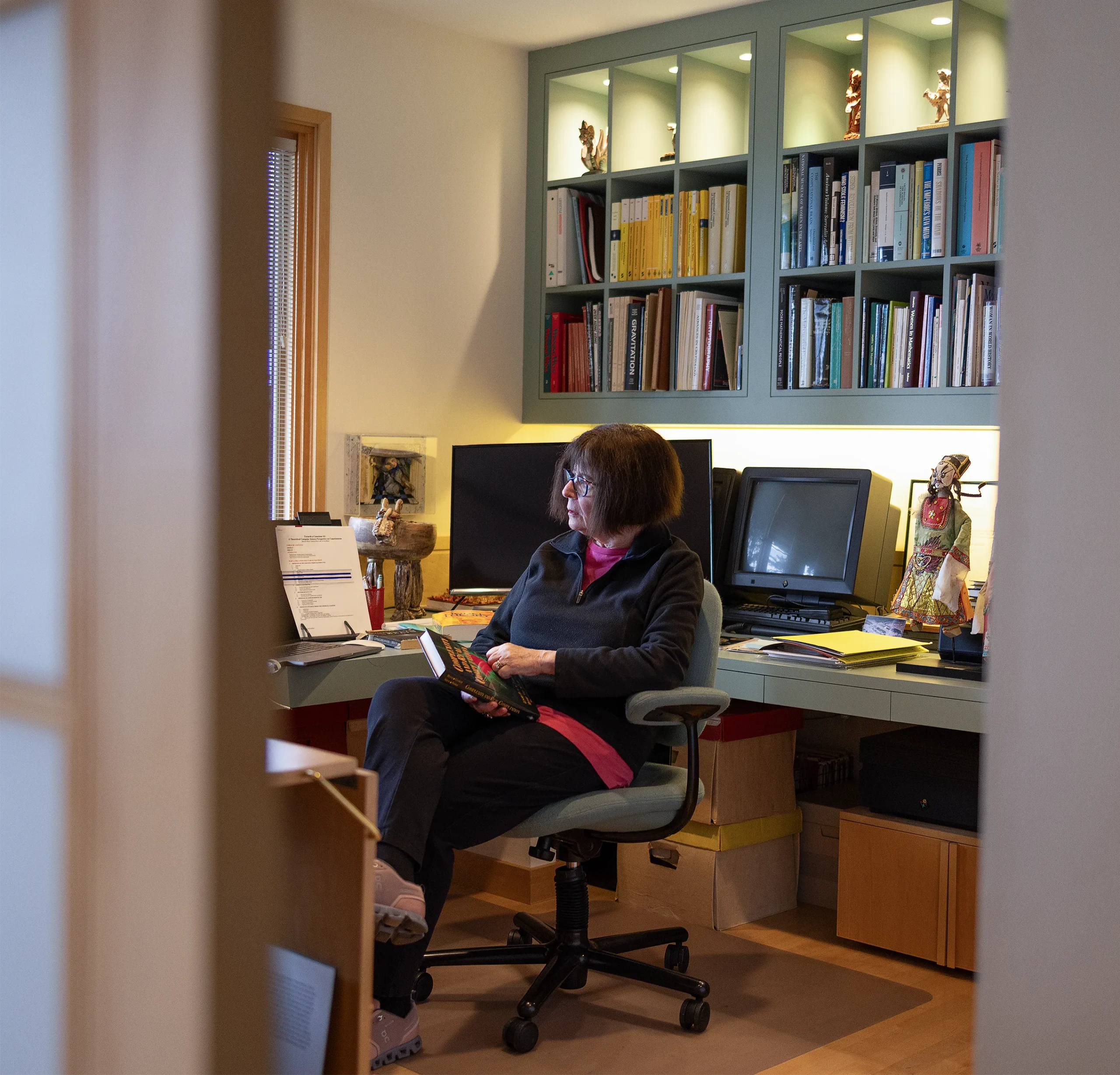 Lenore Blum in a black sweater sits in her office, with books and monitors behind her