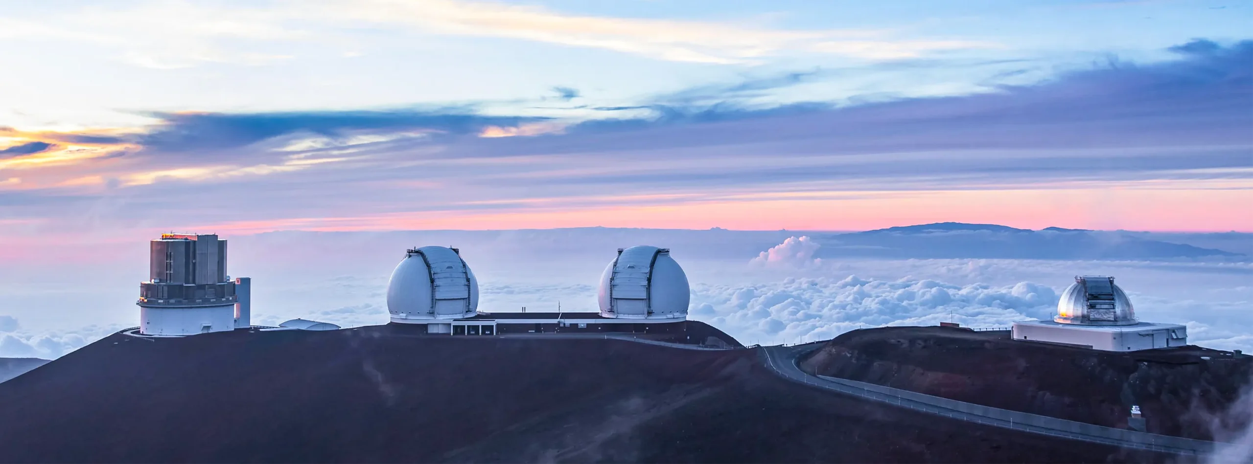 Four telescopes atop Maunakea’s summit. The horizon is pink, and clouds swirl below the summit.