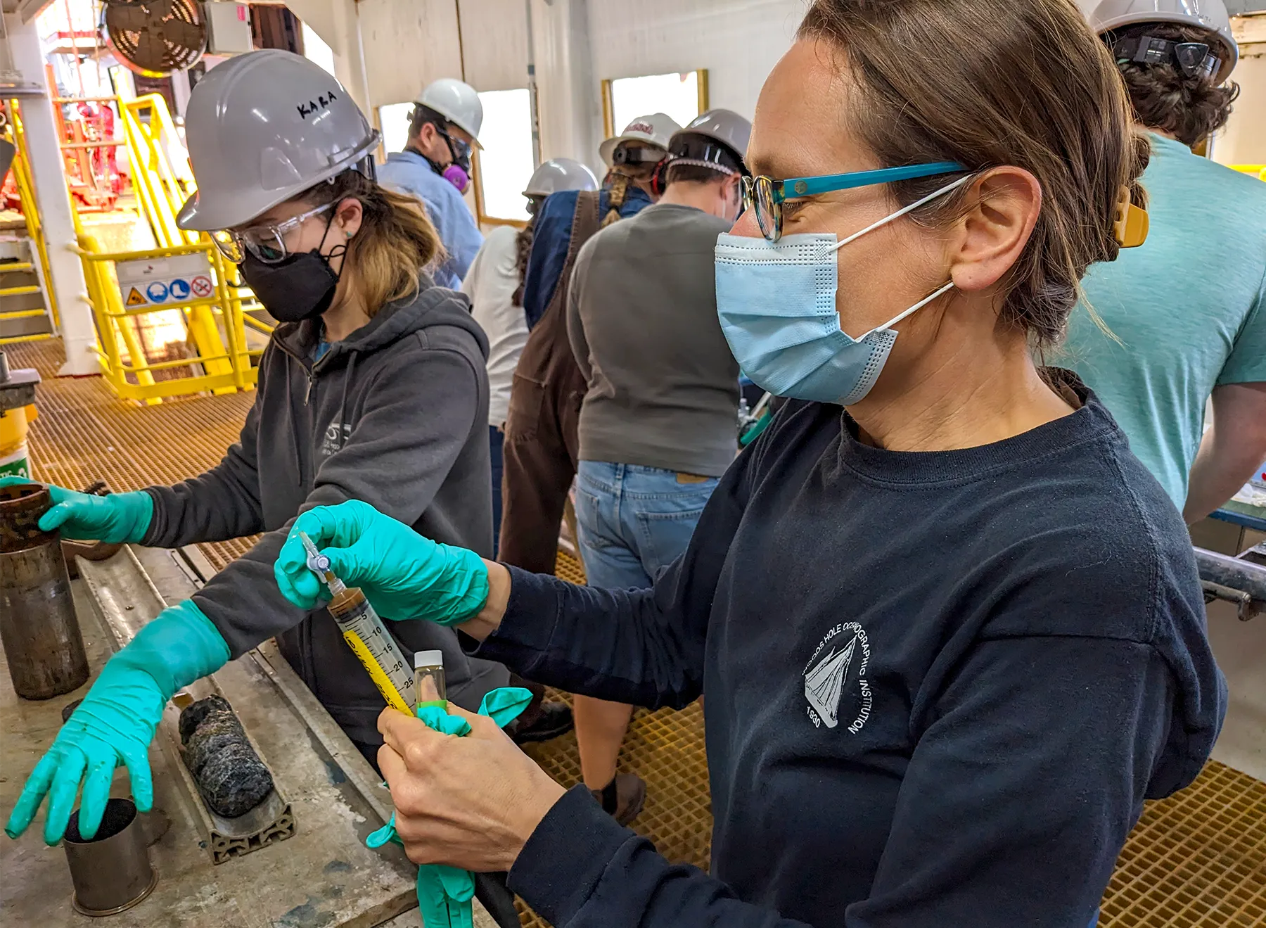 Susan Lang processing samples in a laboratory aboard a ship. She is holding a large syringe and wearing turquoise gloves, a face mask, and a long-sleeved shirt that says “Woods Hole Oceanographic Institution.”