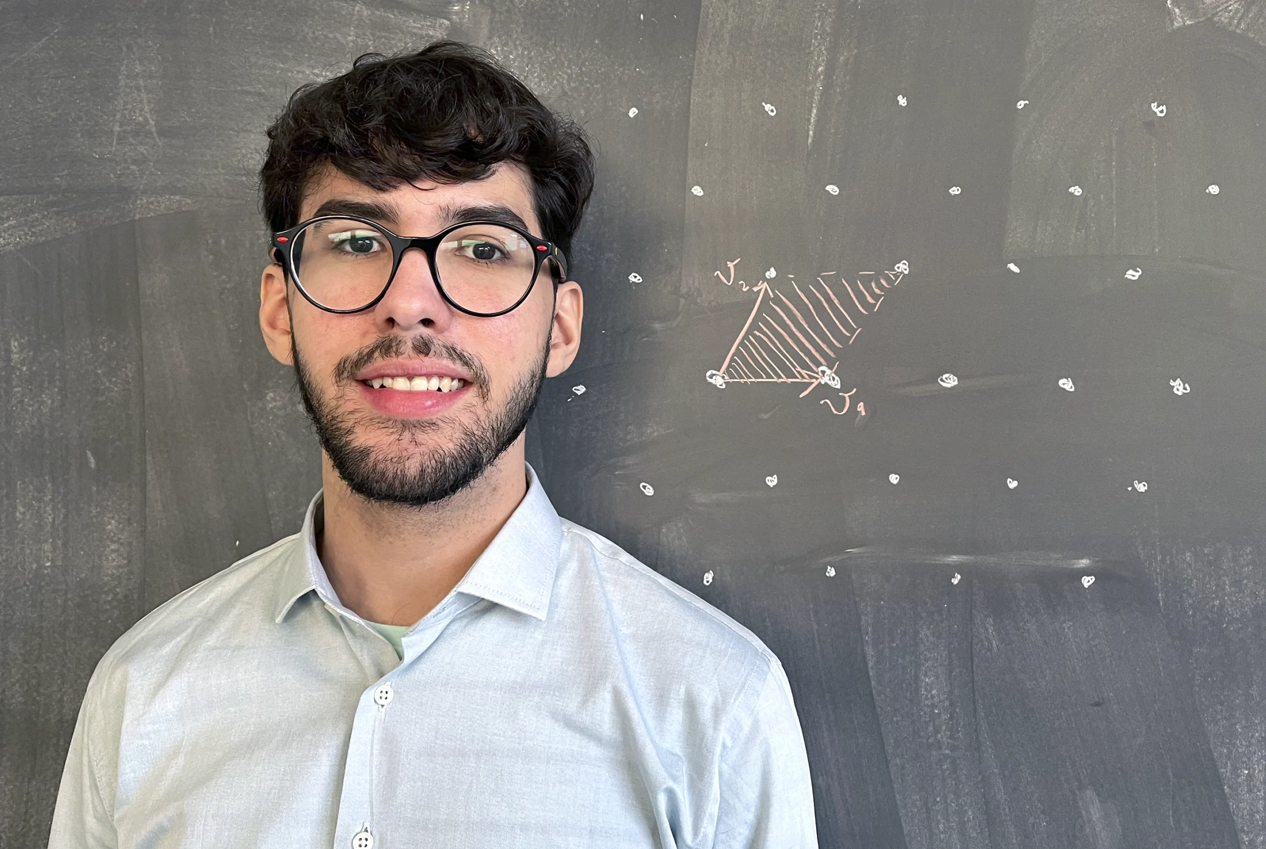 Victor Reis in a light shirt stands in front of a chalkboard with mathematical notation on it.