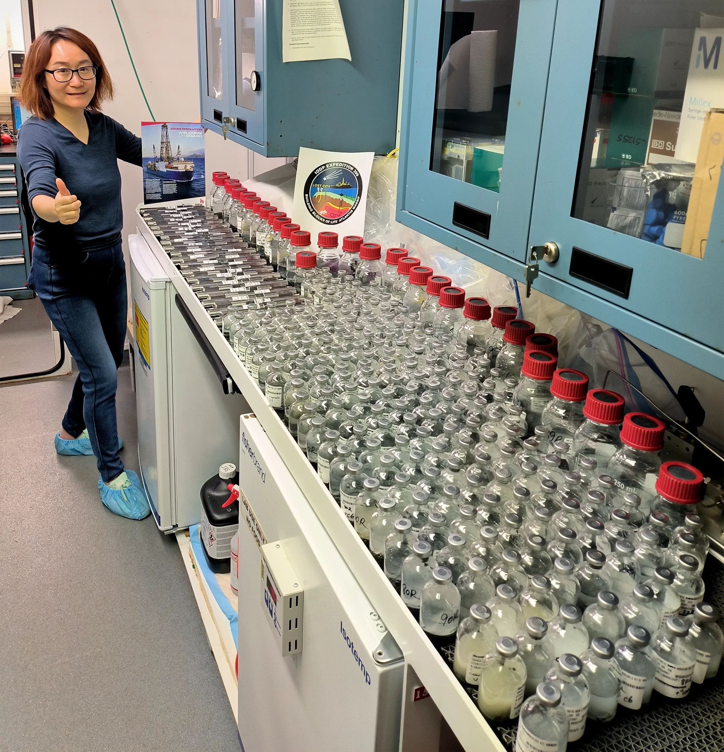 Fengping Wang aboard a ship. She is giving a thumbs-up and smiling while standing next to a table that’s covered in hundreds of glass bottles.