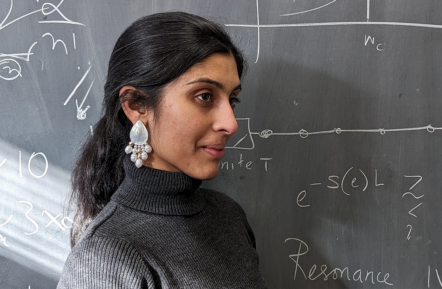 A woman wearing seashell earrings stands in front of a chalkboard depicting a chain of particles.