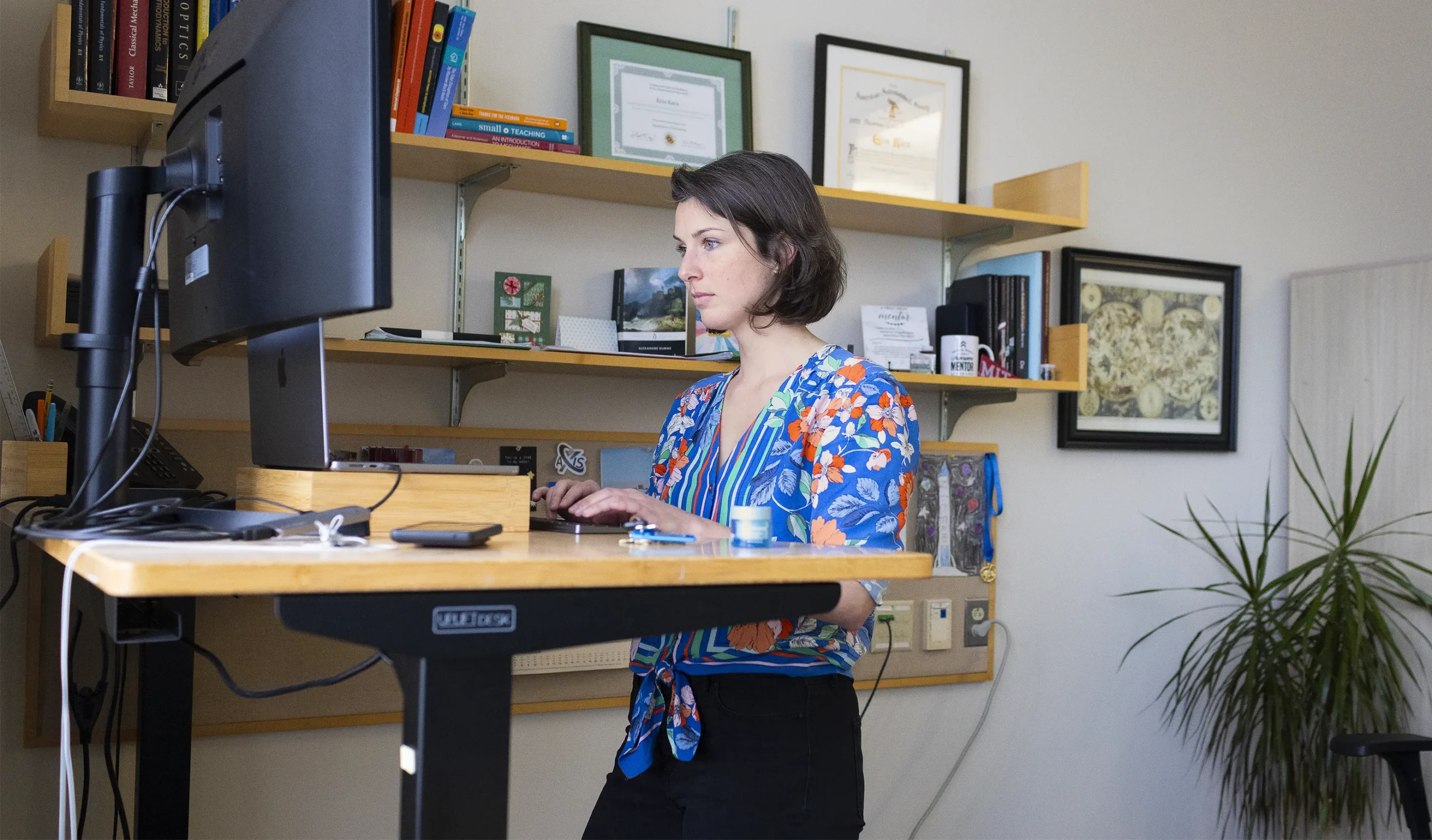 Kara in her office, working on her computer. The shelves behind her are filled with diplomas, books, a drawing and other objects.