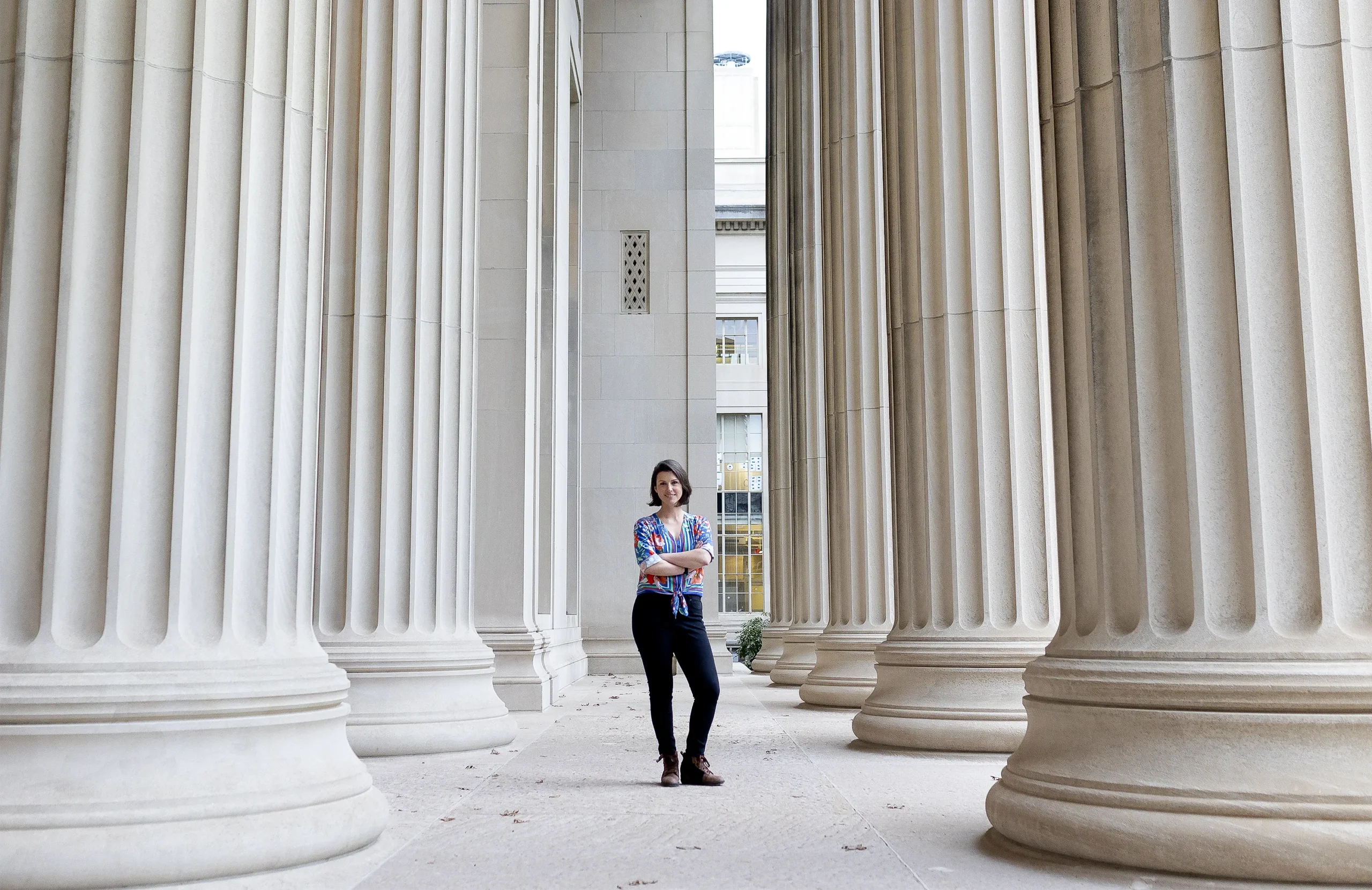 Kara is outside on the MIT campus, standing in a walkway lined by large columns.