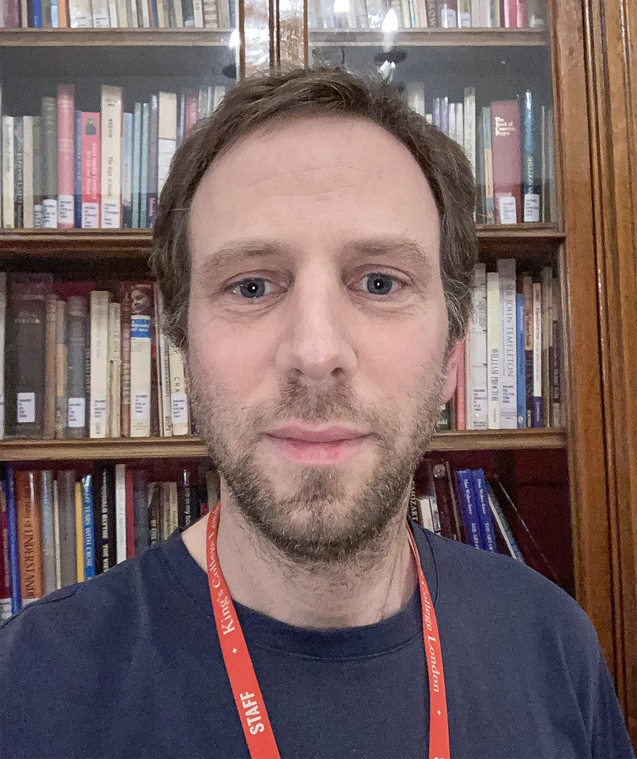 A man wearing a red lanyard stands in front of a bookshelf.