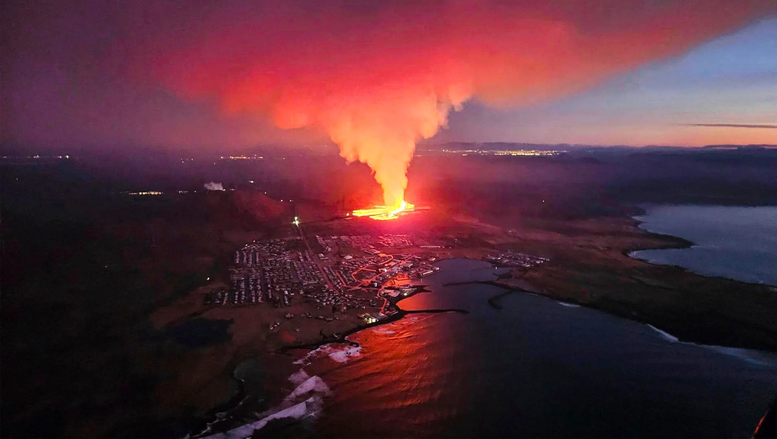 Bird’s-eye view of a fiery eruption filling the sky with smoke and ash next to a coastal town.