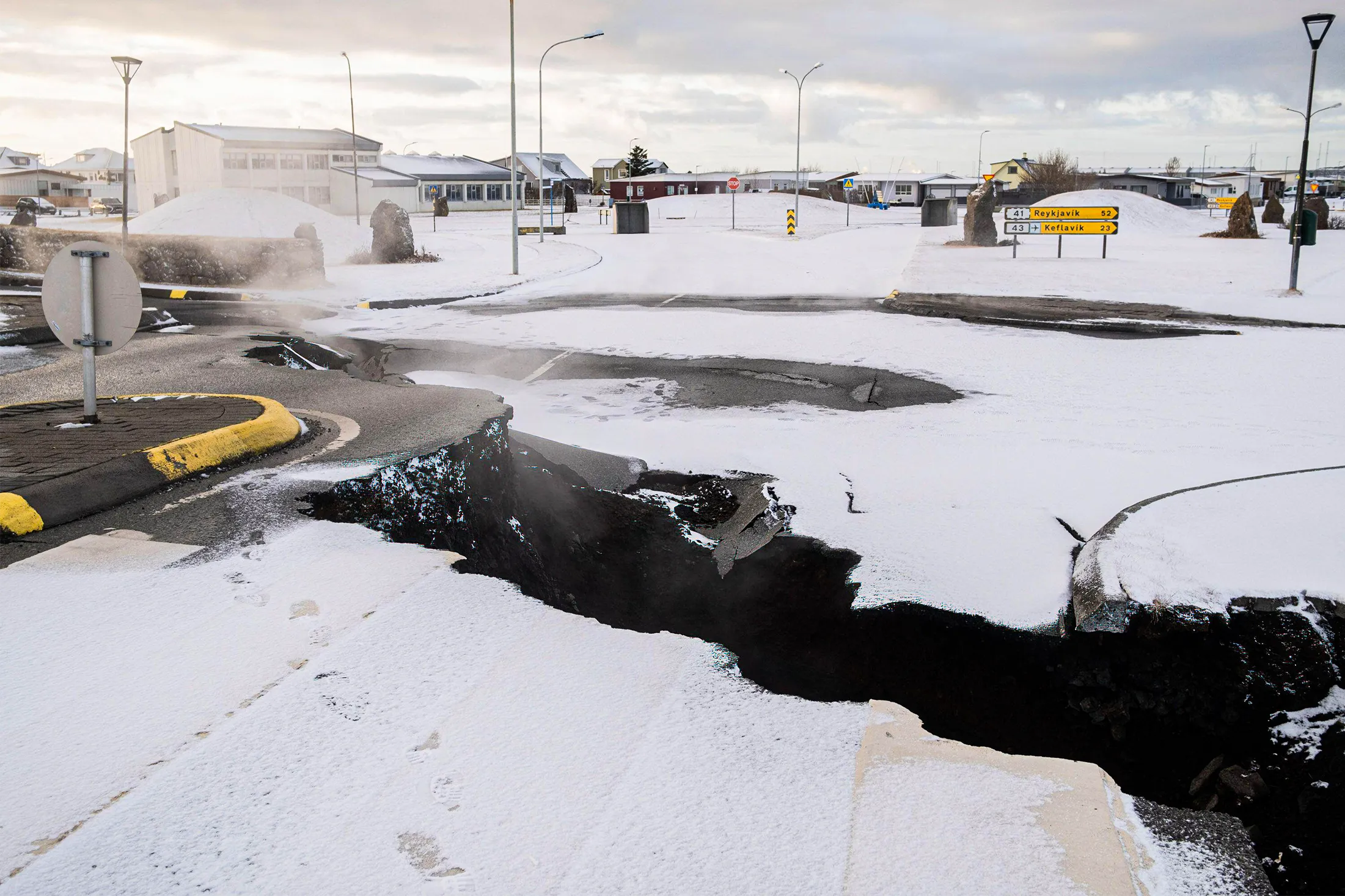 A crack in Earth’s surface crosses a city street in a snowy scene with buildings and streetlights in the background.