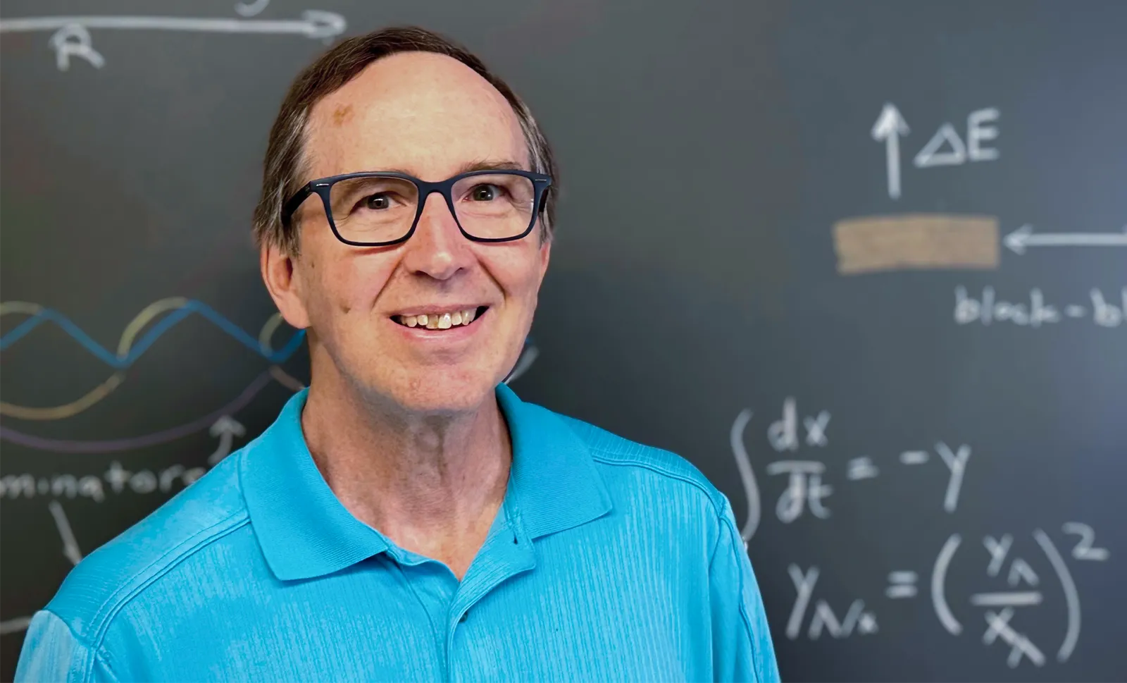 A man in a blue shirt stands in front of a chalkboard.