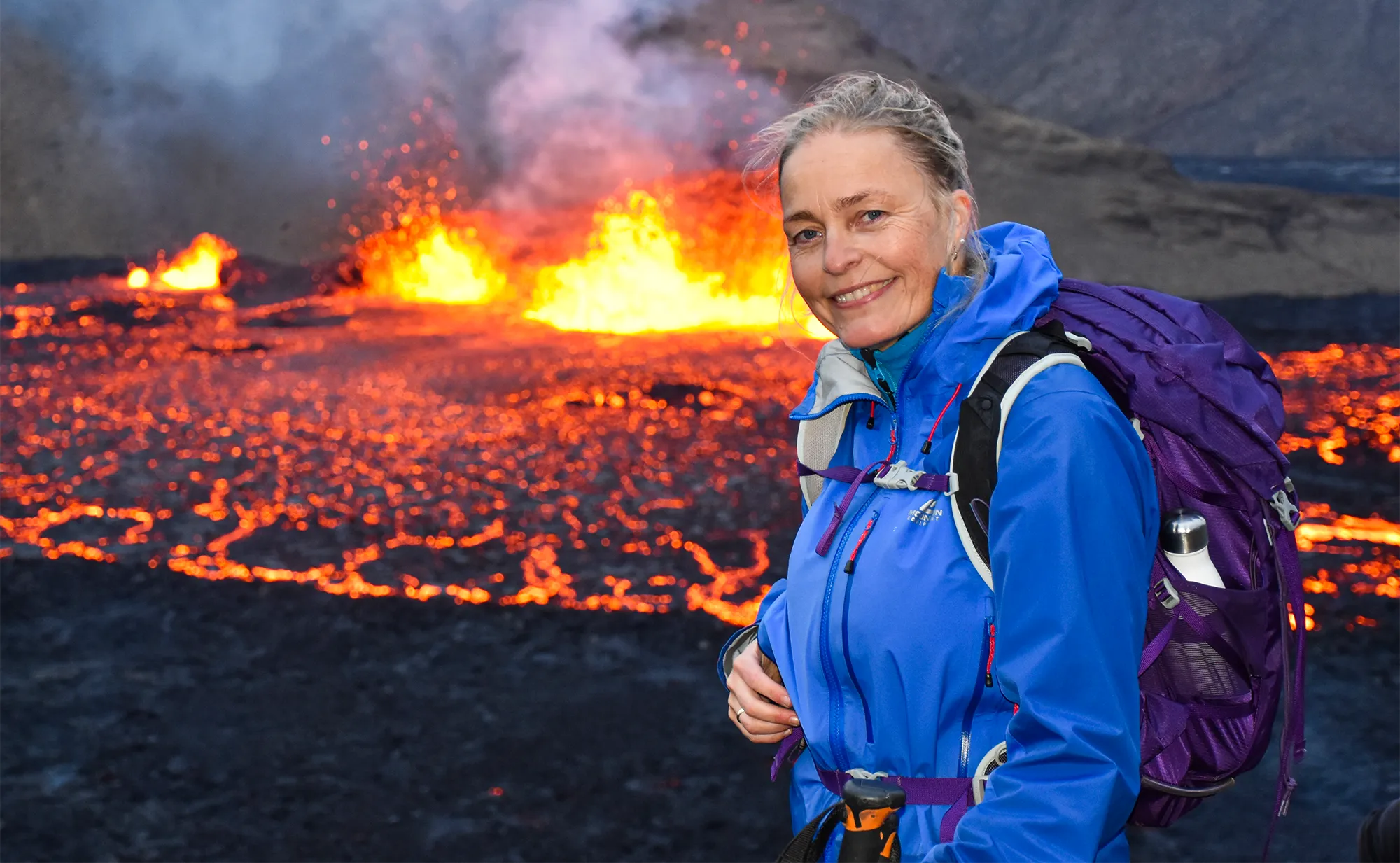 A smiling woman in a blue winter jacket stands in front of a fiery lava flow.