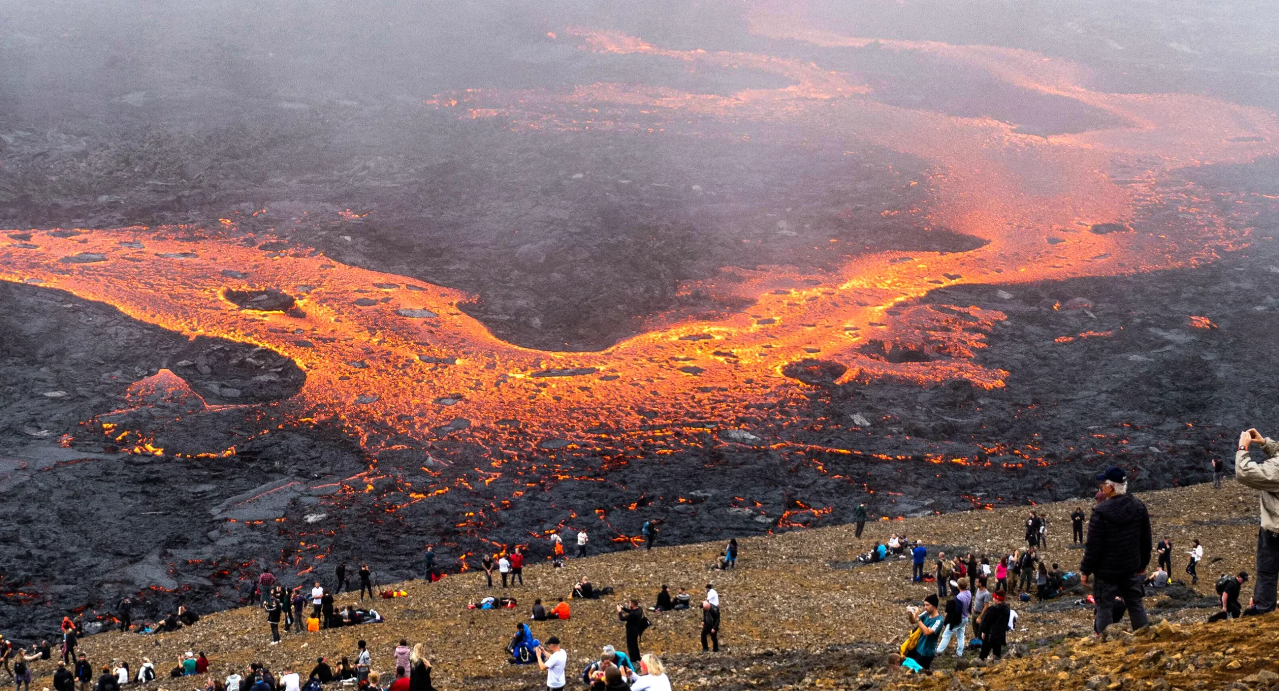 People sitting on a grassy hill in the foreground look out over a valley full of glowing rivers of lava.