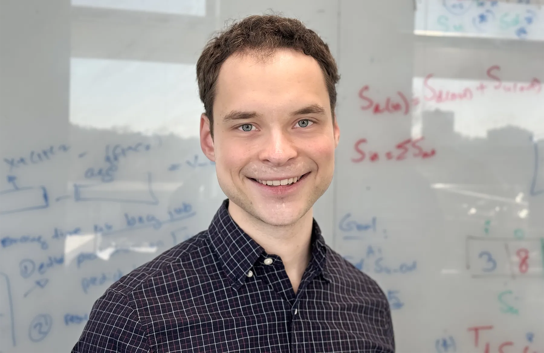 William Kuszmaul in a checkered shirt stands in front of a whiteboard with figures on it.