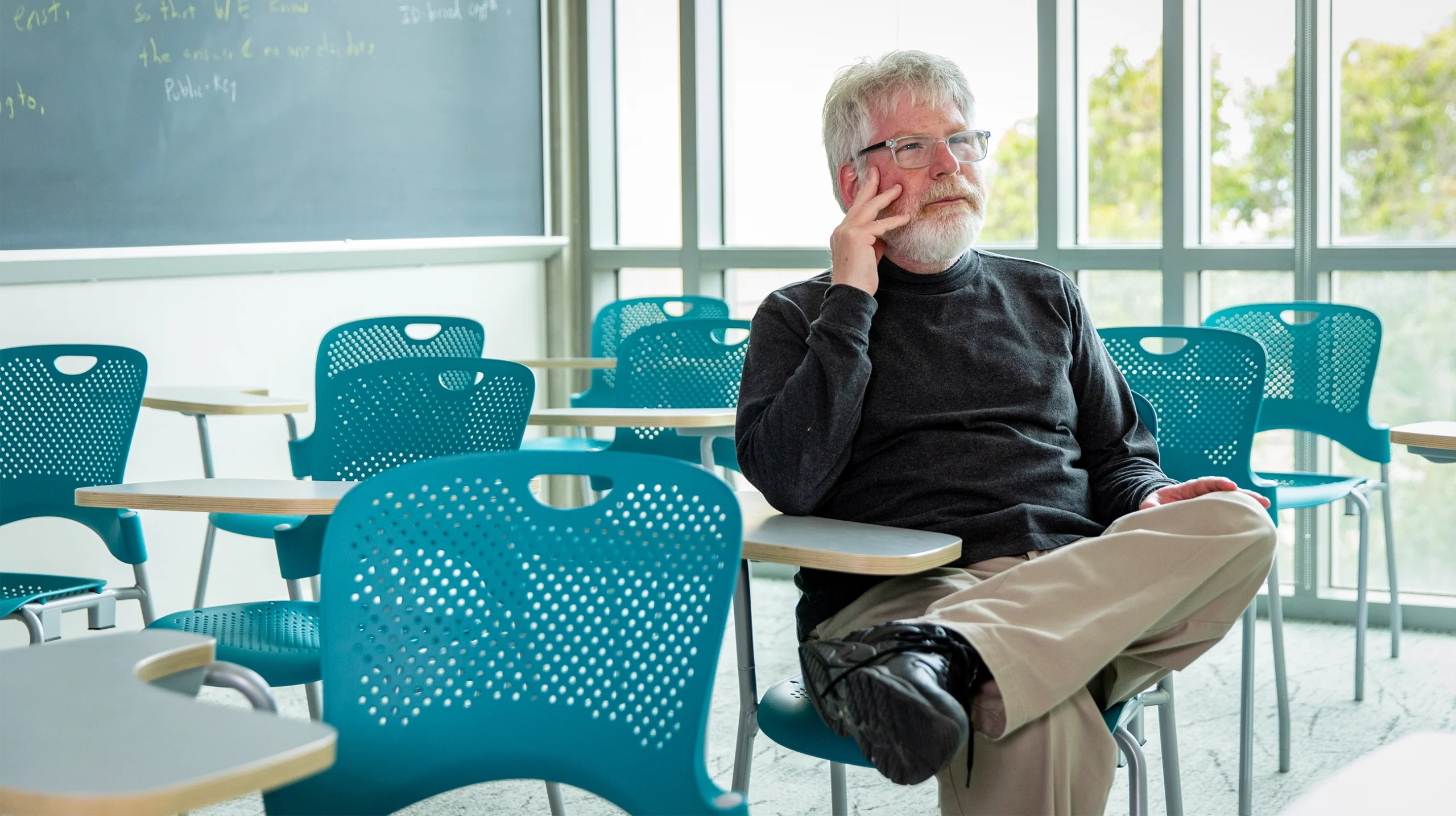 Russell Impagliazzo, in khakis and a dark shirt, sits at a desk in a classroom