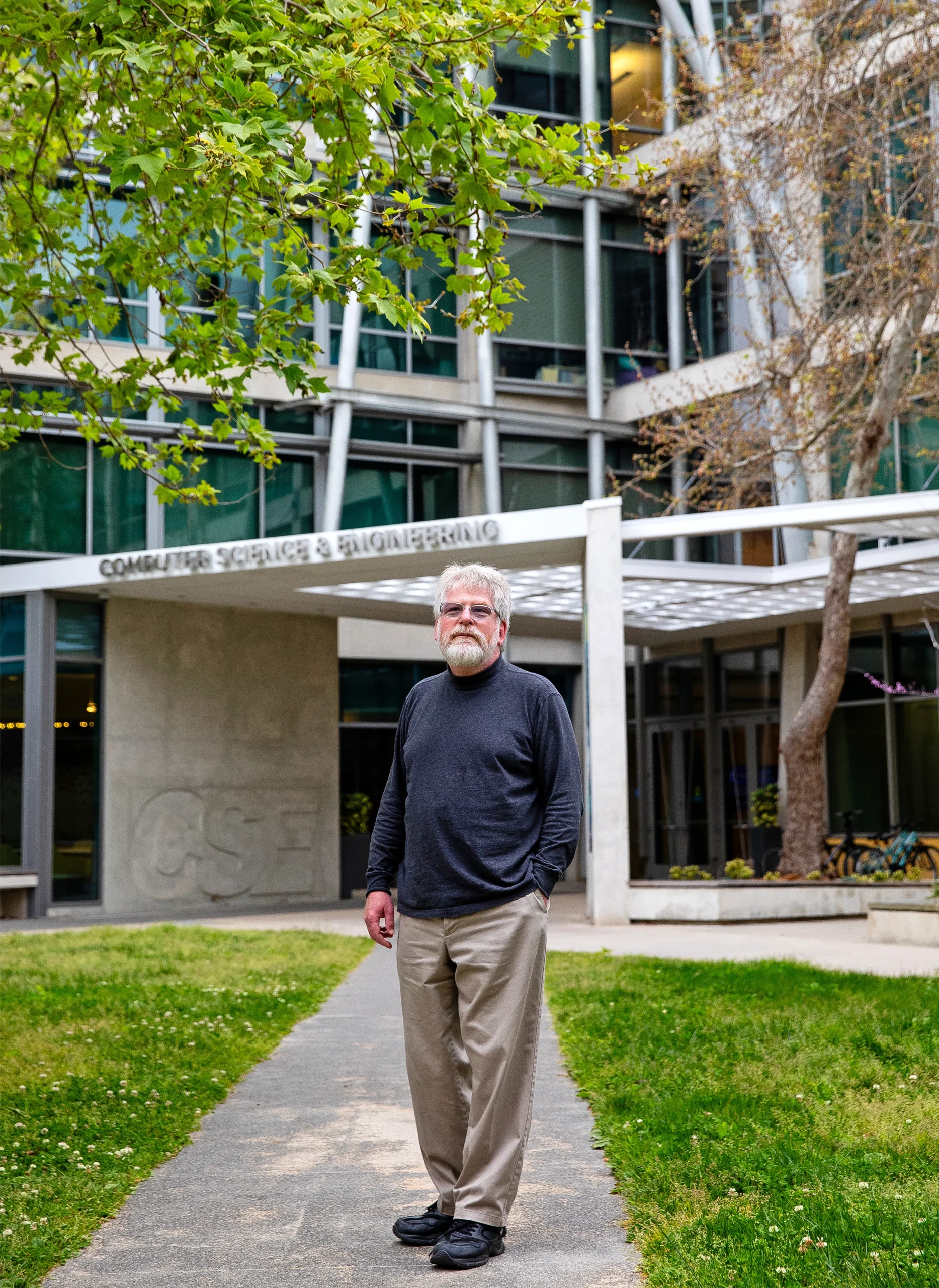 Russell Impagliazzo, in khakis and a dark shirt, stands on a path outside a building