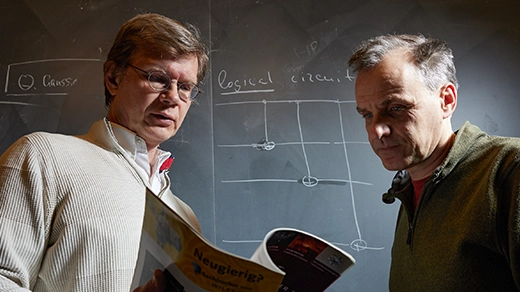 Two men standing in front of a blackboard are glancing down at the open page of a journal that the man on the left is holding. The blackboard features a sketch labeled “logical circuit.”