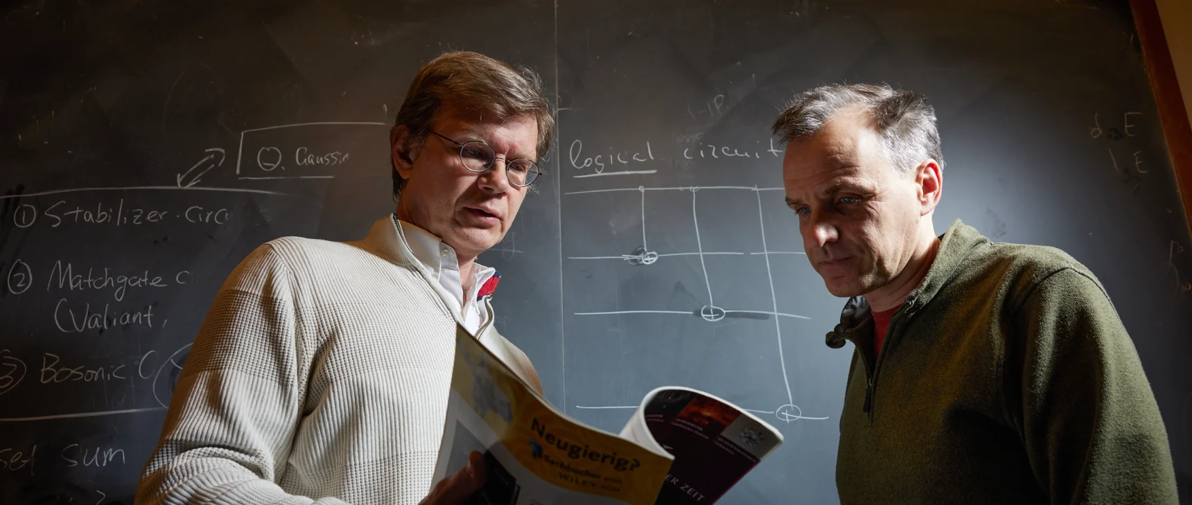 Two men standing in front of a blackboard are glancing down at the open page of a journal that the man on the left is holding. The blackboard features a sketch labeled “logical circuit.”