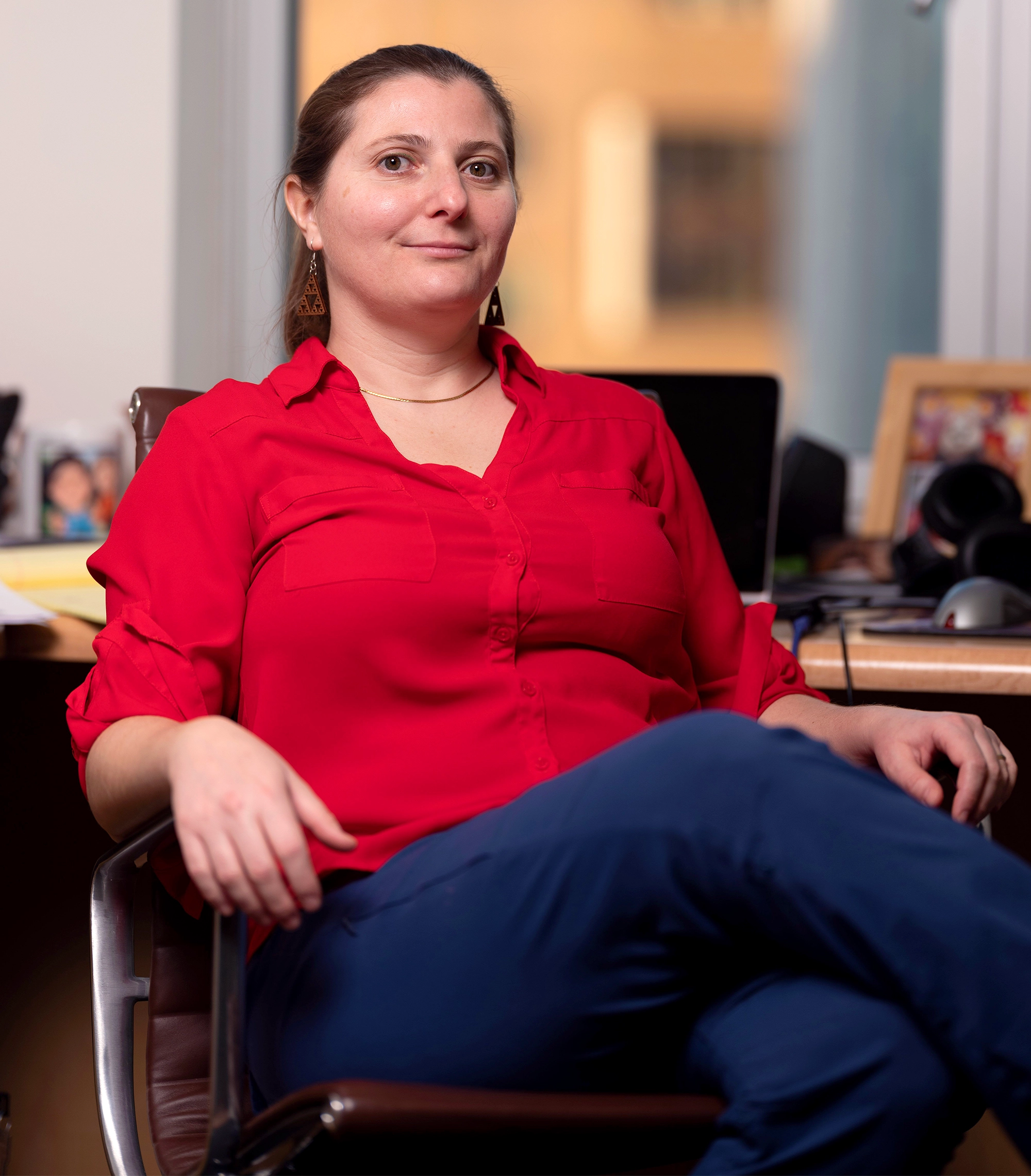 Virginia Vassilevska Williams in a red shirt and blue pants sits with her back to a desk and window.