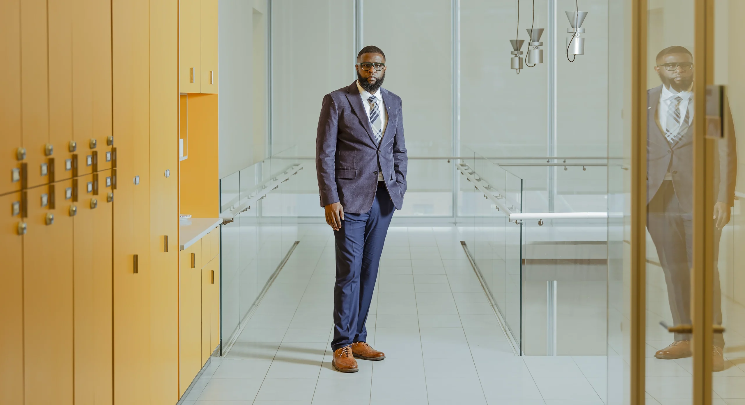 The neuroscientist Ishmail Abdus-Saboor, wearing a jacket and tie, stands in a hallway near an atrium, flanked by orange lockers and glass partitions.