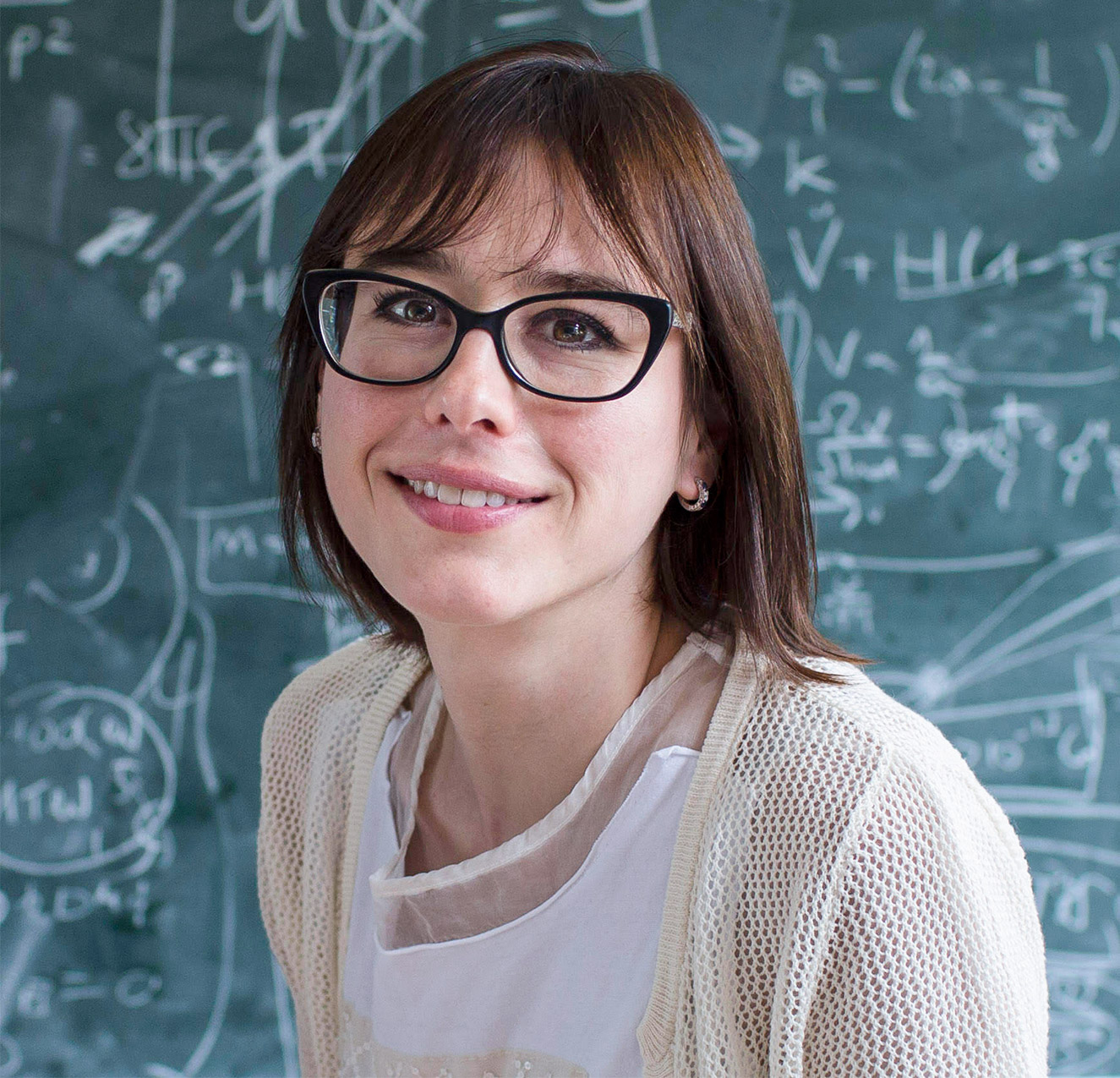 Portrait of a woman with brown hair and glasses smiling while seated in front of a blackboard.