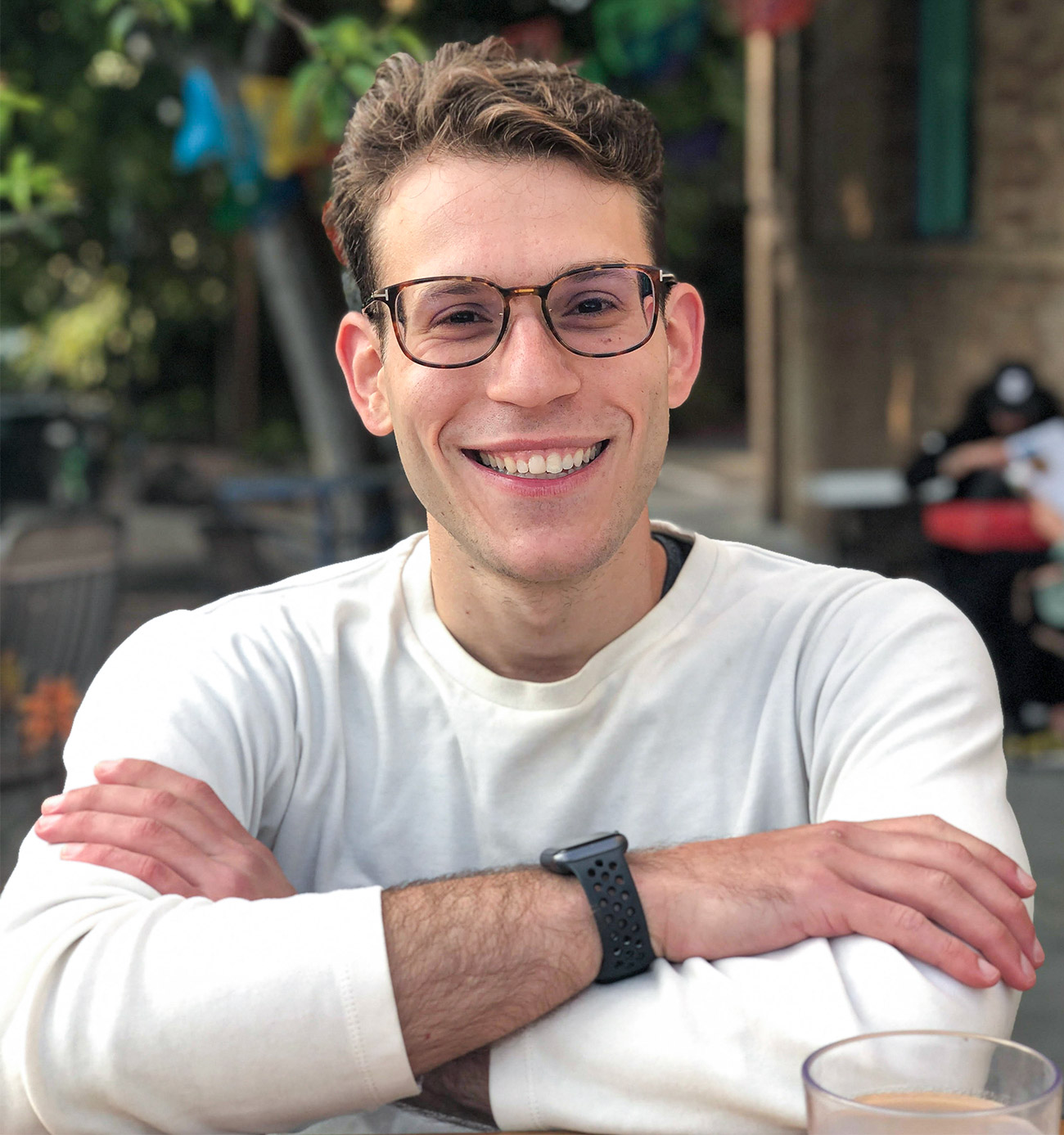 A man in a white shirt and glasses seated at an outdoor table smiles at the camera.