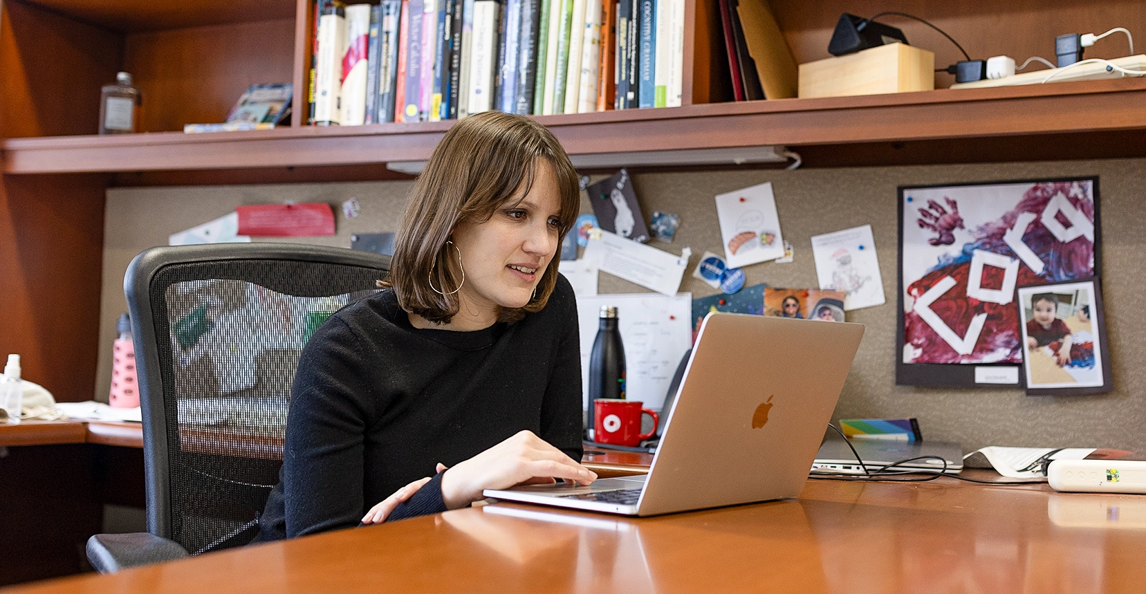 Ellie Pavlick in a black shirt sits at a desk with a laptop in front of a shelf full of books