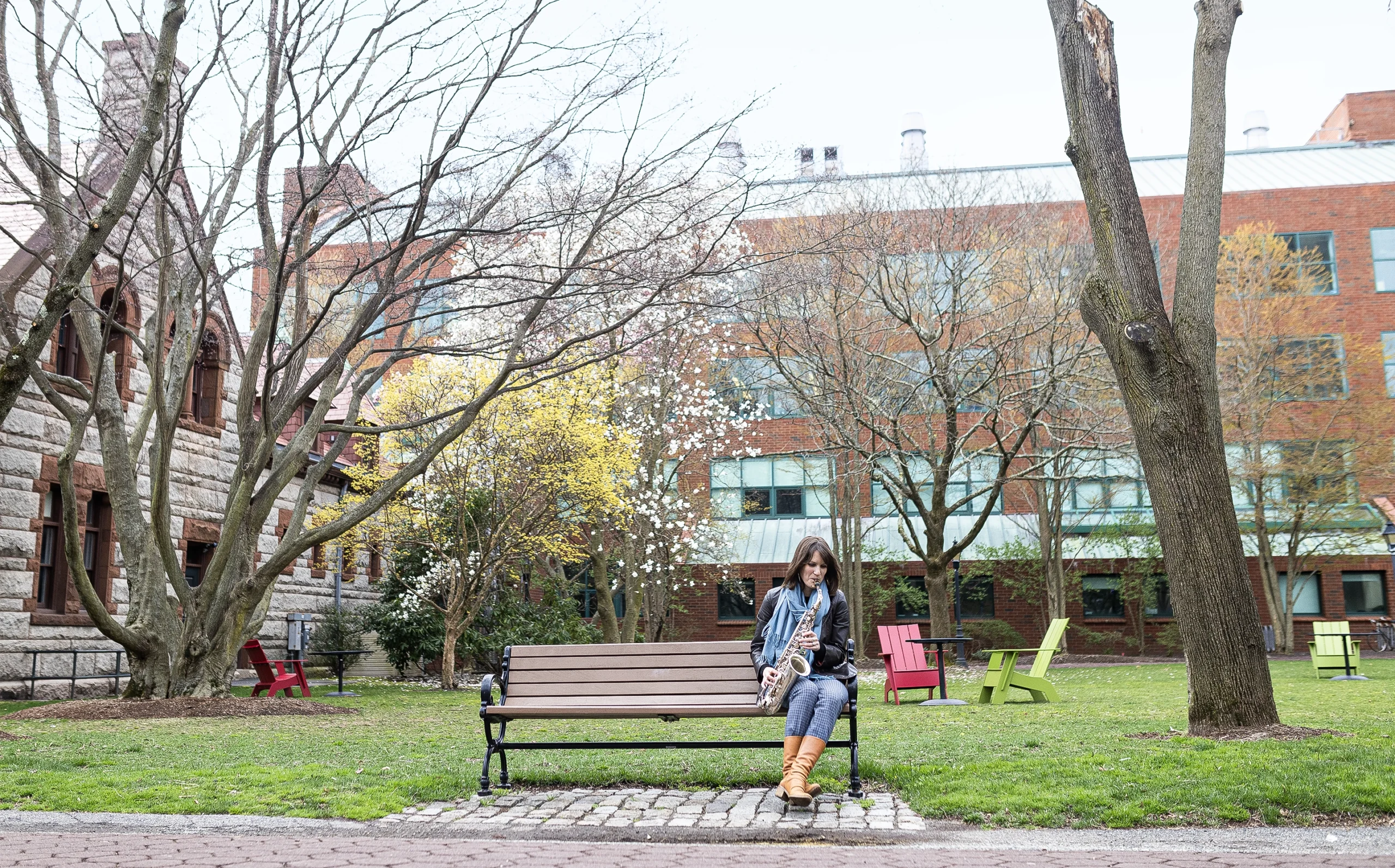 Ellie Pavlick sits on the edge of a bench playing a saxophone in a grassy square