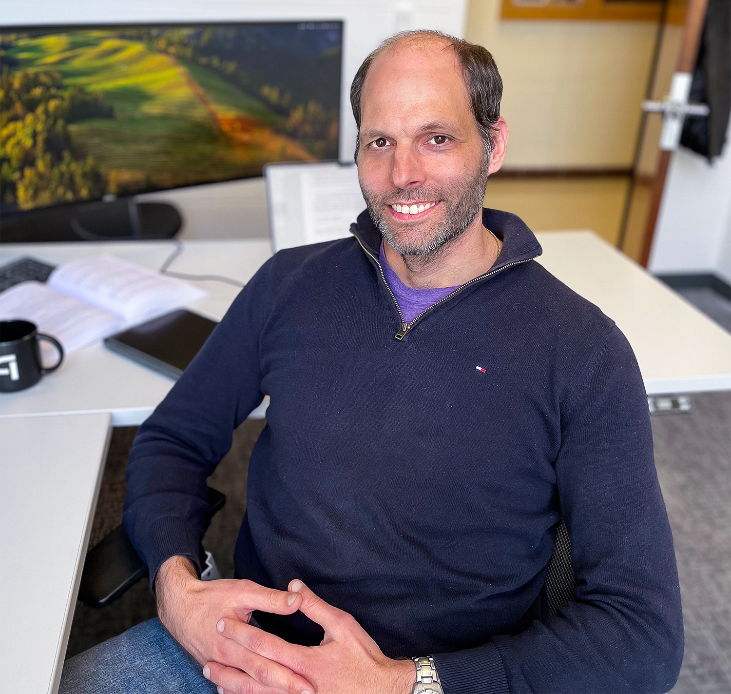 A smiling man sits at a desk with a double-width monitor in the background.