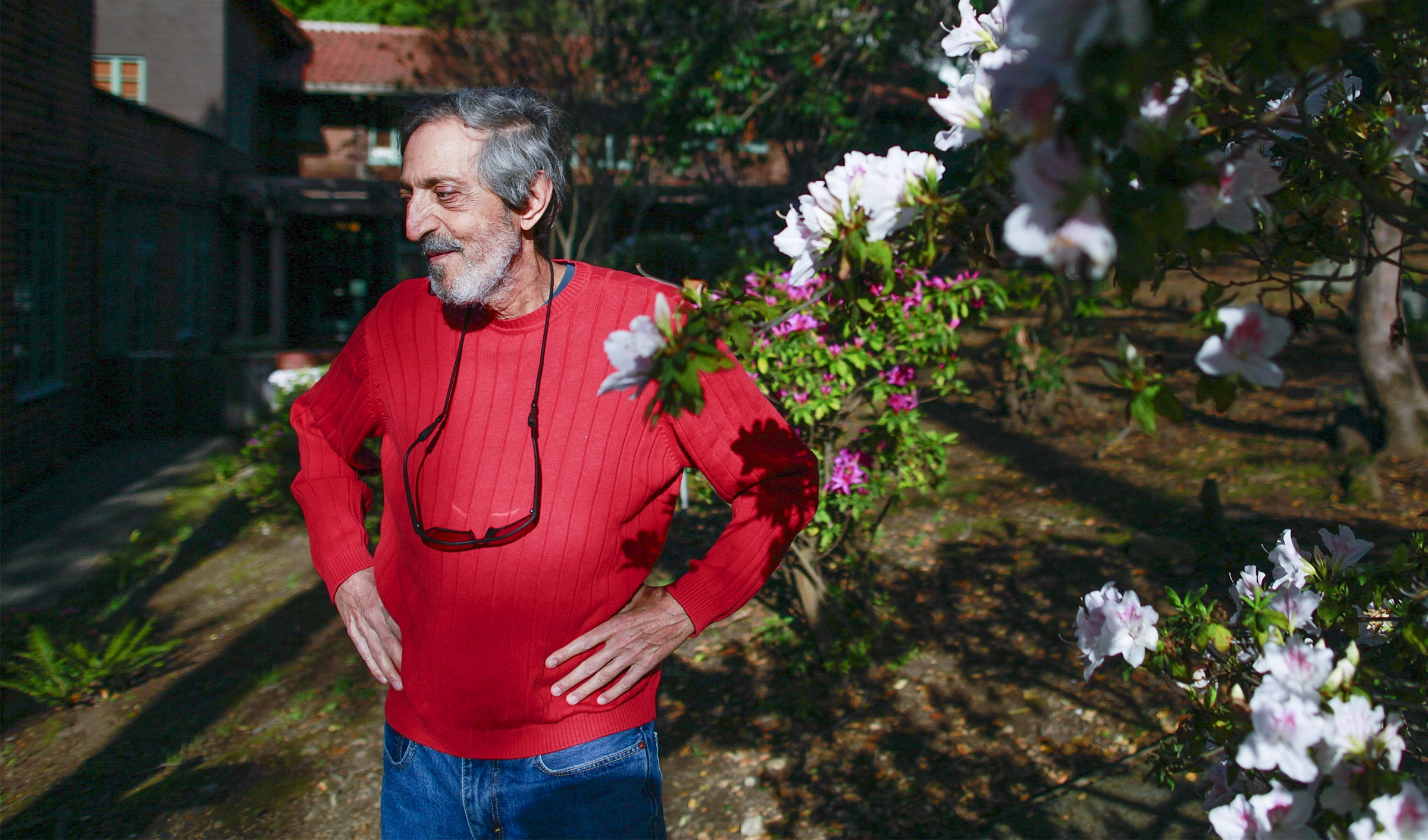 Avi Wigderson in a red sweater and blue jeans stands outside by white and pink flowers.