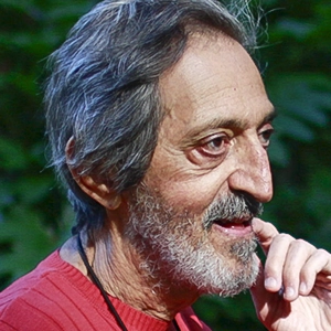 Avi Wigderson in a red sweater stands in front of rocks and a tree.